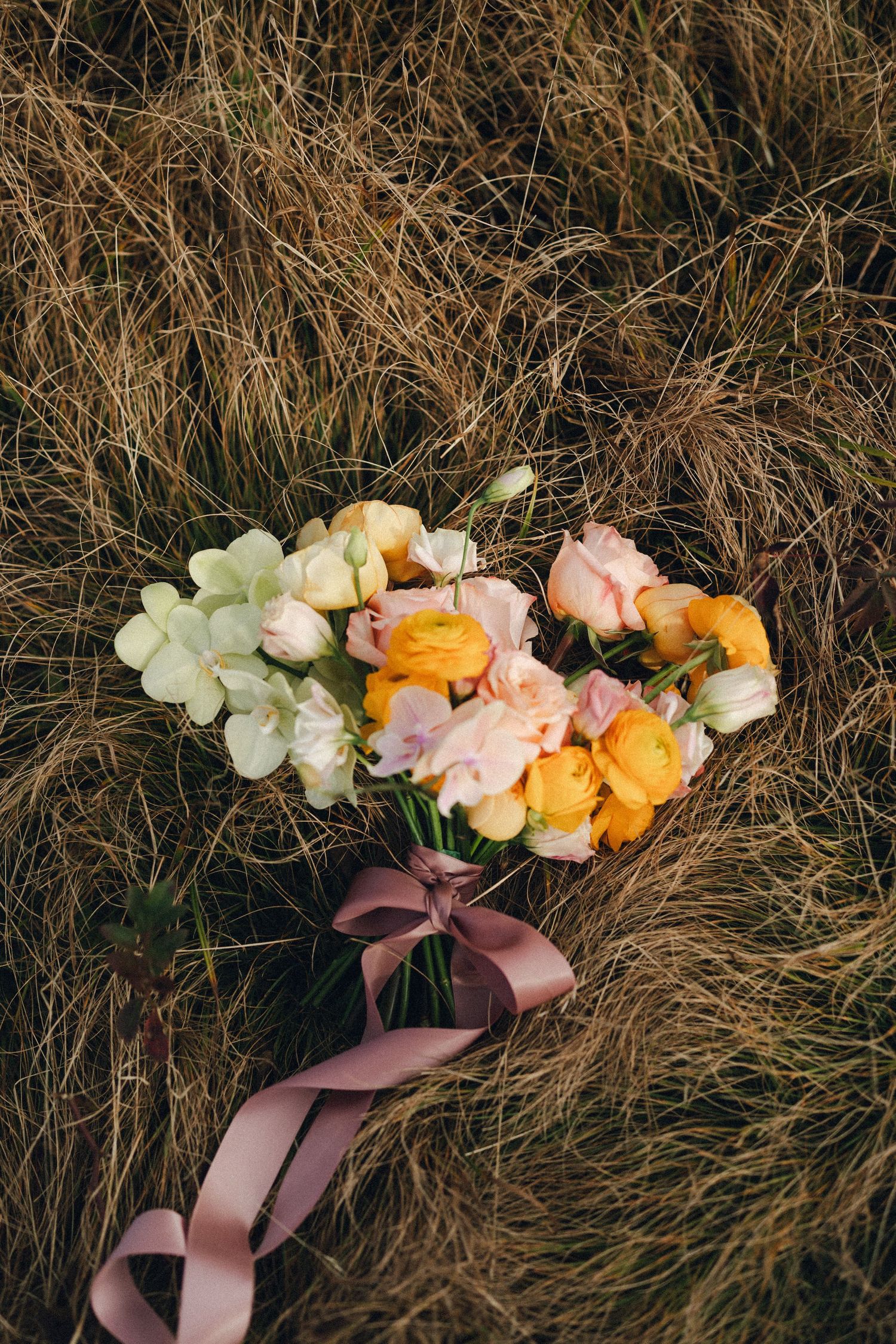 Colorful bridal bouquet of orange ranunculus, pink roses, and green hydrangea resting on dry golden grass.