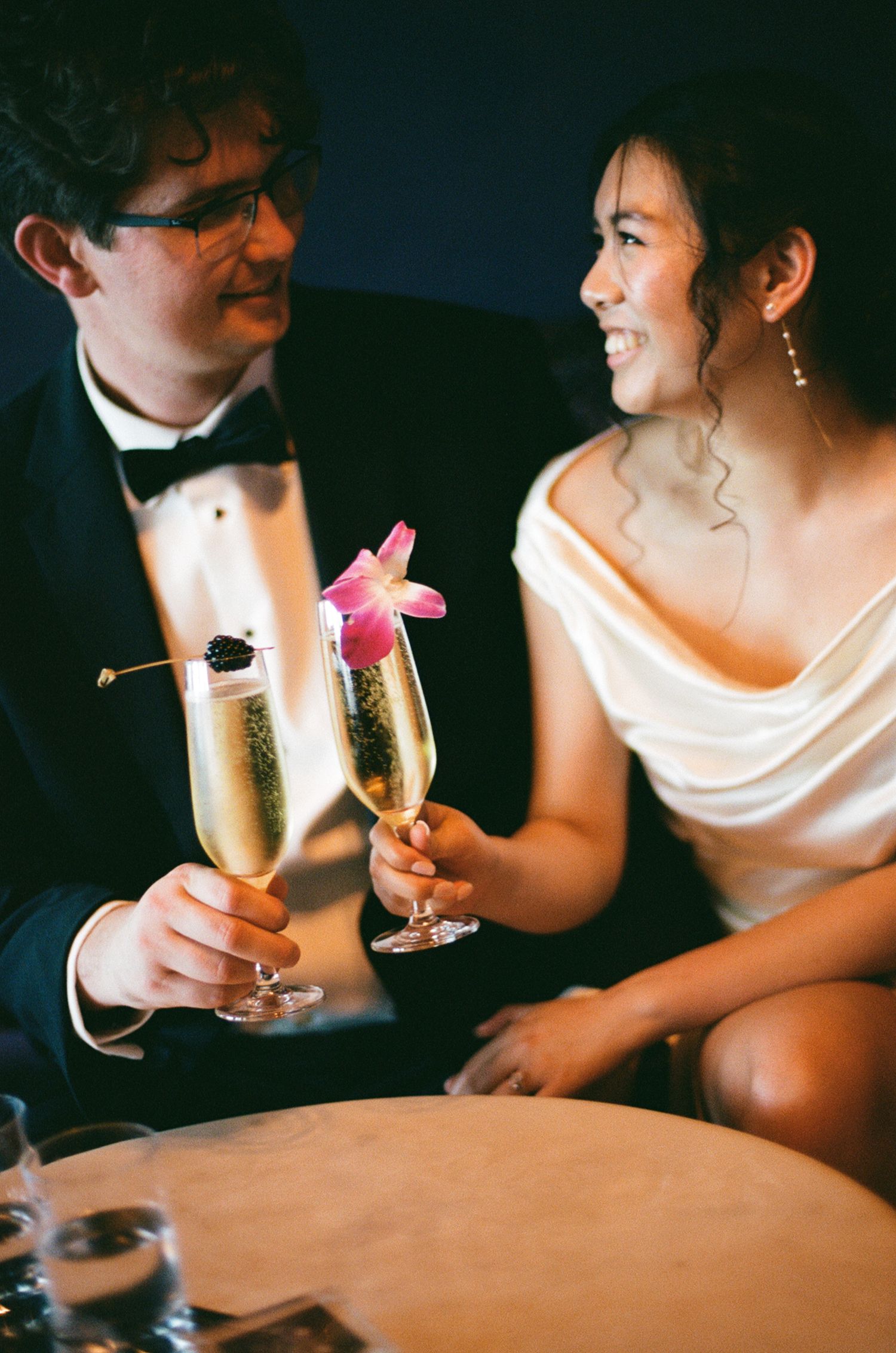 Bride and groom toast with champagne flutes garnished with pink flowers at a wedding.