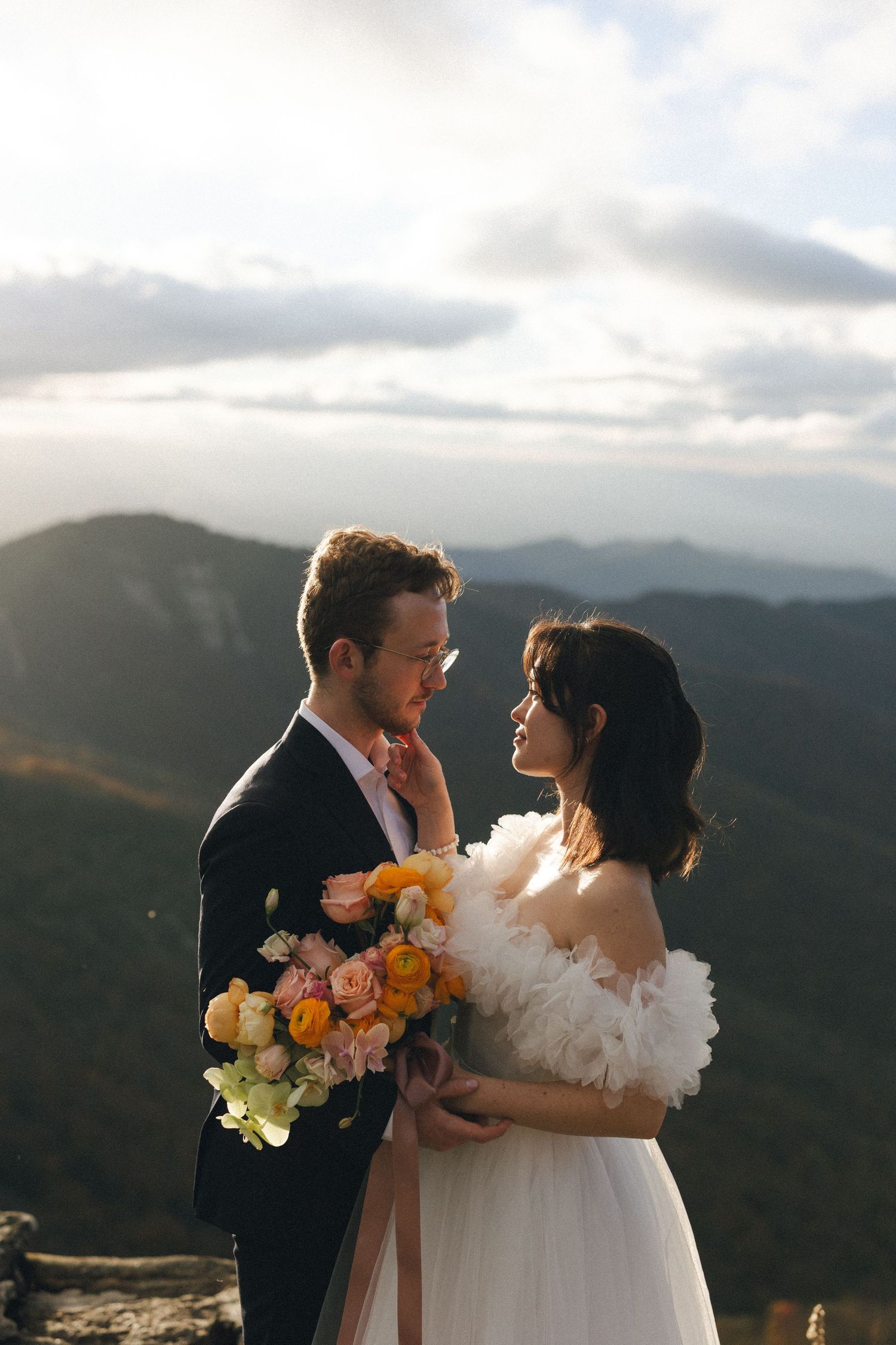 Bride and groom gazing at each other in the blue ridge mountains, bride holding a vibrant orange and pink arrangement.