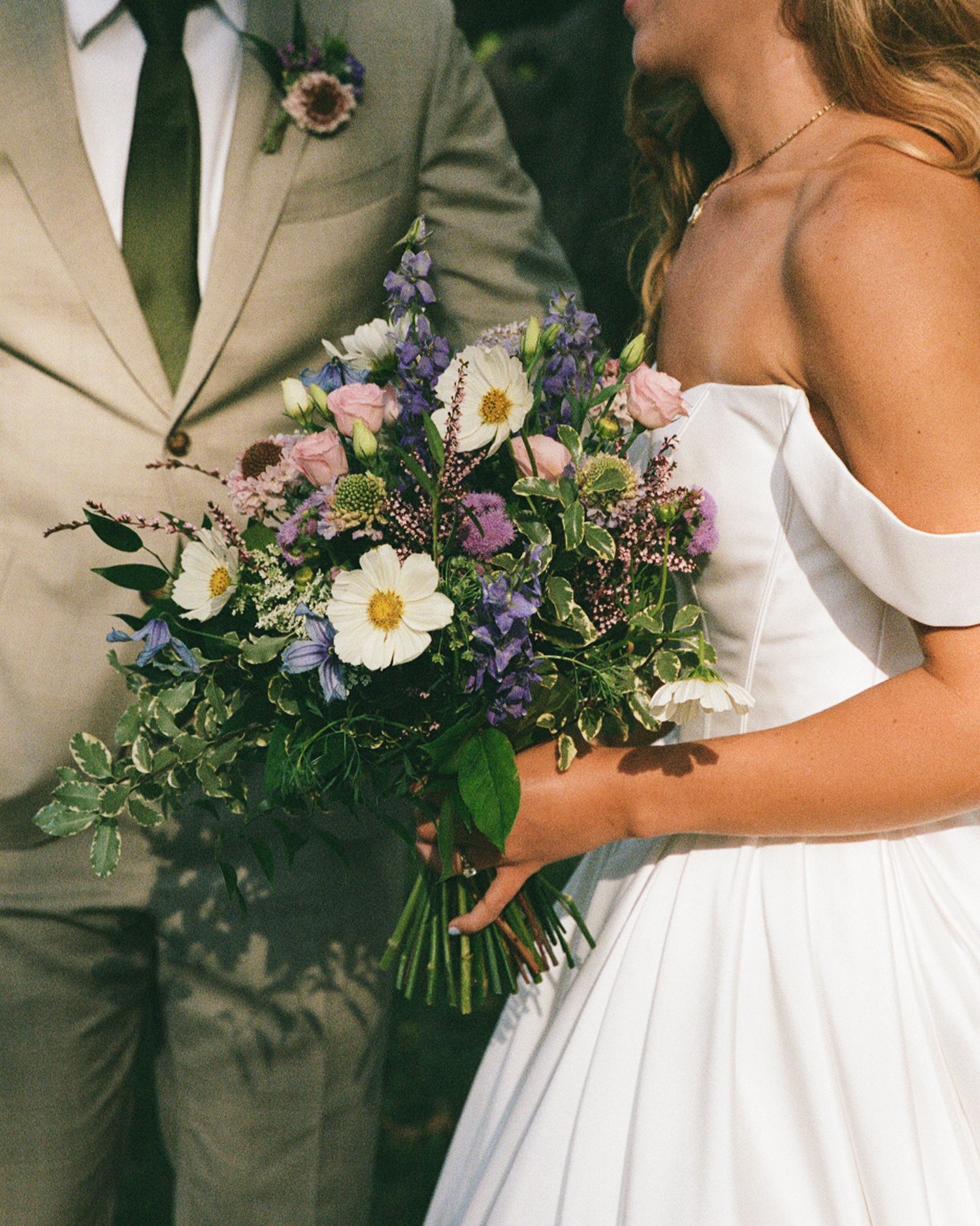 Bride holding lush wildflower bouquet with white cosmos, purple lavender, and greenery beside groom in tan suit.