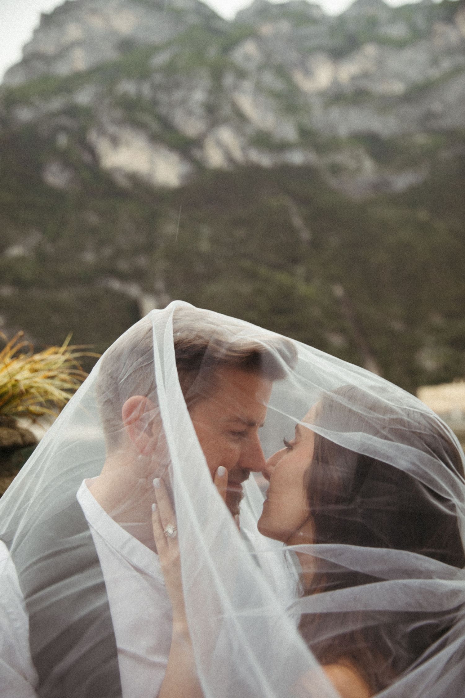 Romantic wedding couple sharing a kiss under the bride's flowing veil with dramatic mountain scenery behind them.