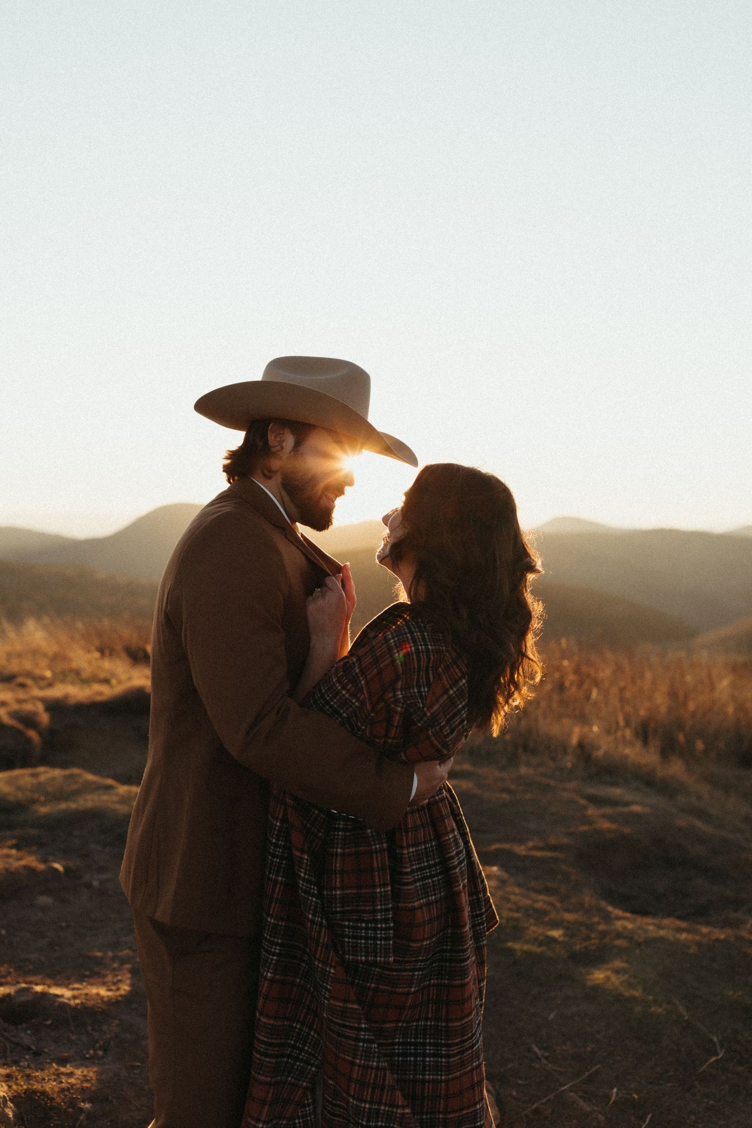 Couple embraces at golden sunset in mountains, man in cowboy hat holds woman in plaid dress.