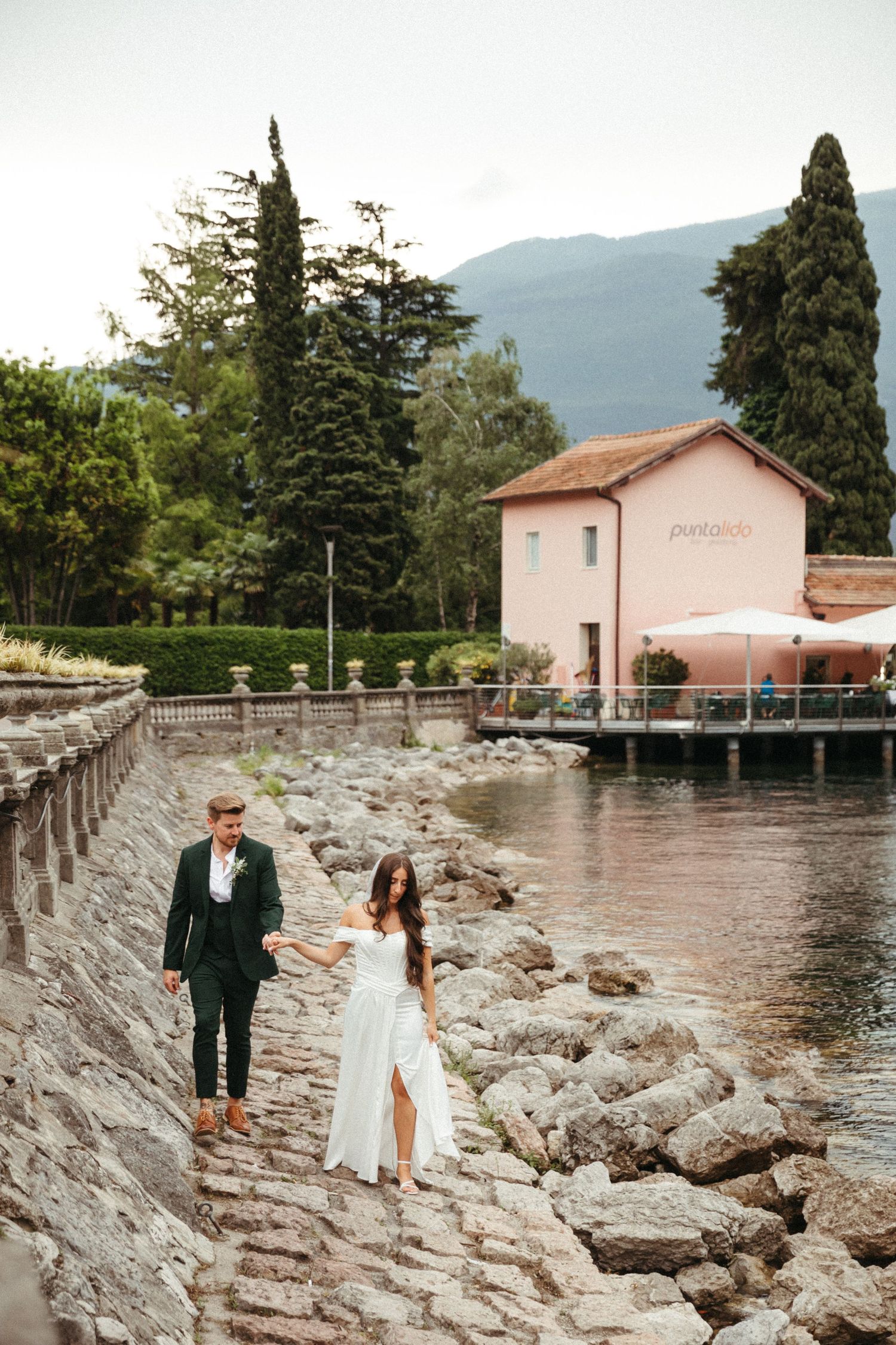 Bride and groom walking hand in hand along a rocky lakeside path with a pink building and mountains in the background.