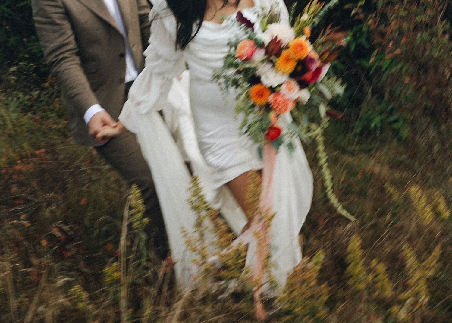 Bride in white dress holding colorful bouquet walking with groom through wildflowers.