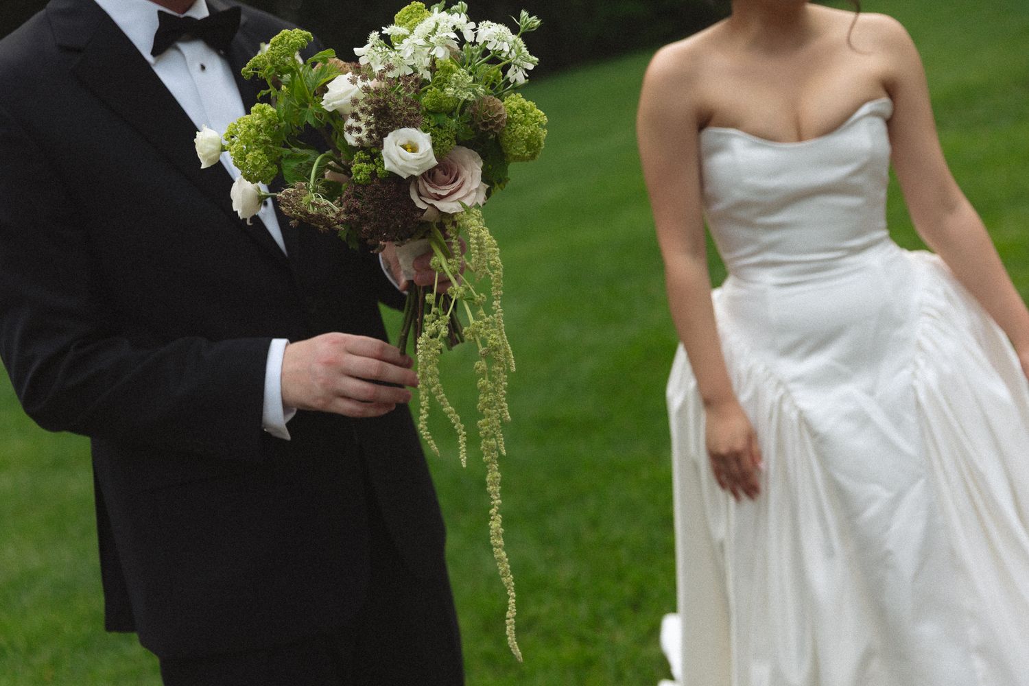 Groom in black tuxedo holds lush green and white bouquet beside bride in strapless white gown.