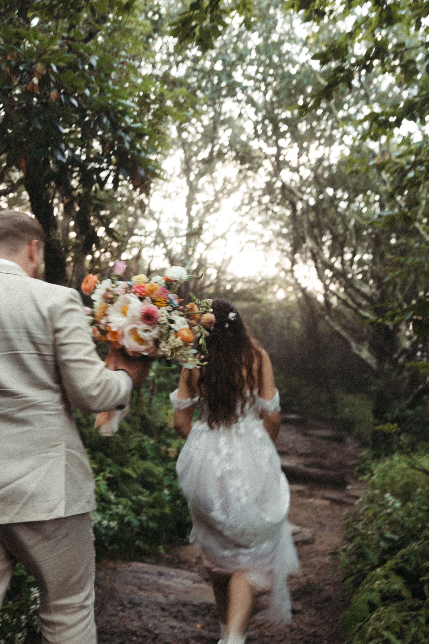 Bride and groom walking hand in hand through sunlit forest path holding colorful bouquet.