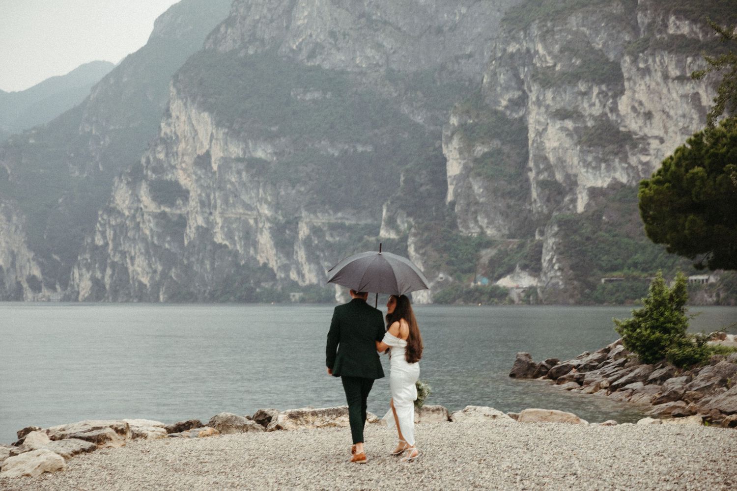 Newlyweds walk along a rocky lakeside shore under an umbrella with dramatic mountain backdrop.