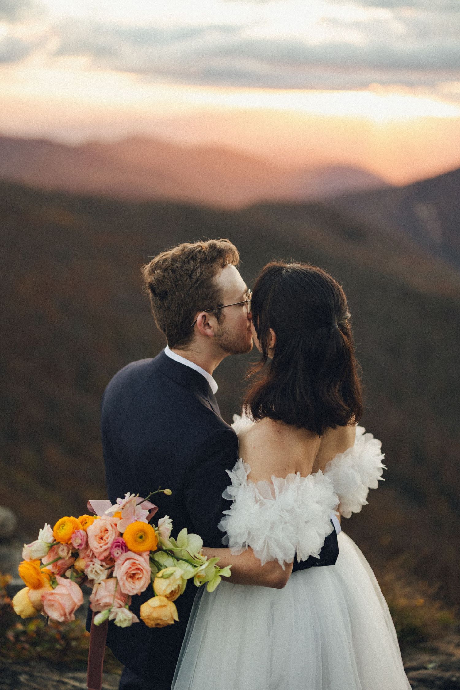 Bride and groom share a romantic kiss at sunset with colorful mountain views and a vibrant floral bouquet.