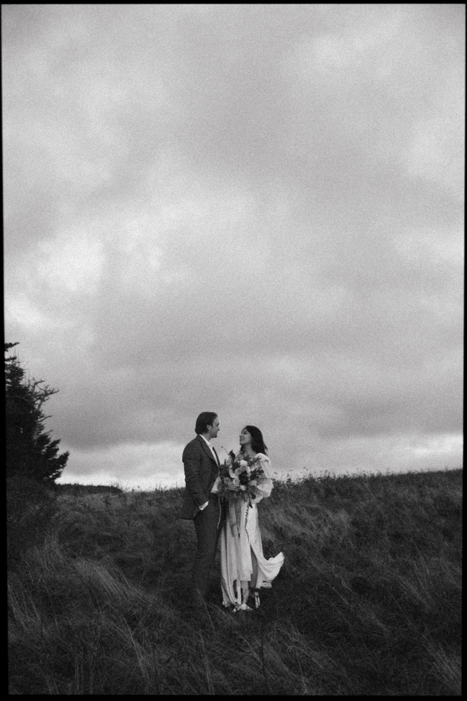 Black and white photo of a couple standing in an open field holding a bouquet under a dramatic cloudy sky.