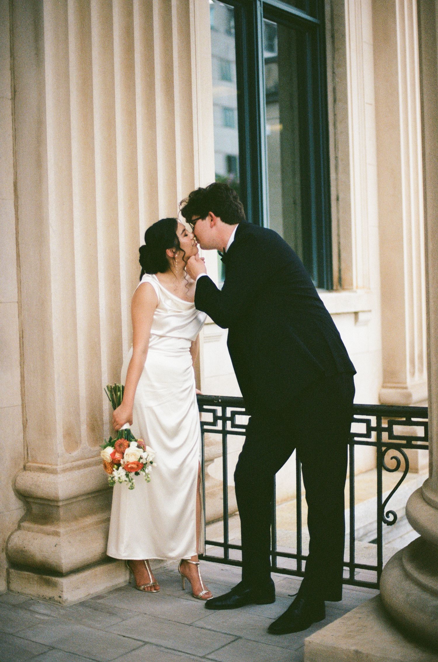 Groom kisses bride beside grand stone columns as she holds a vibrant bridal bouquet.