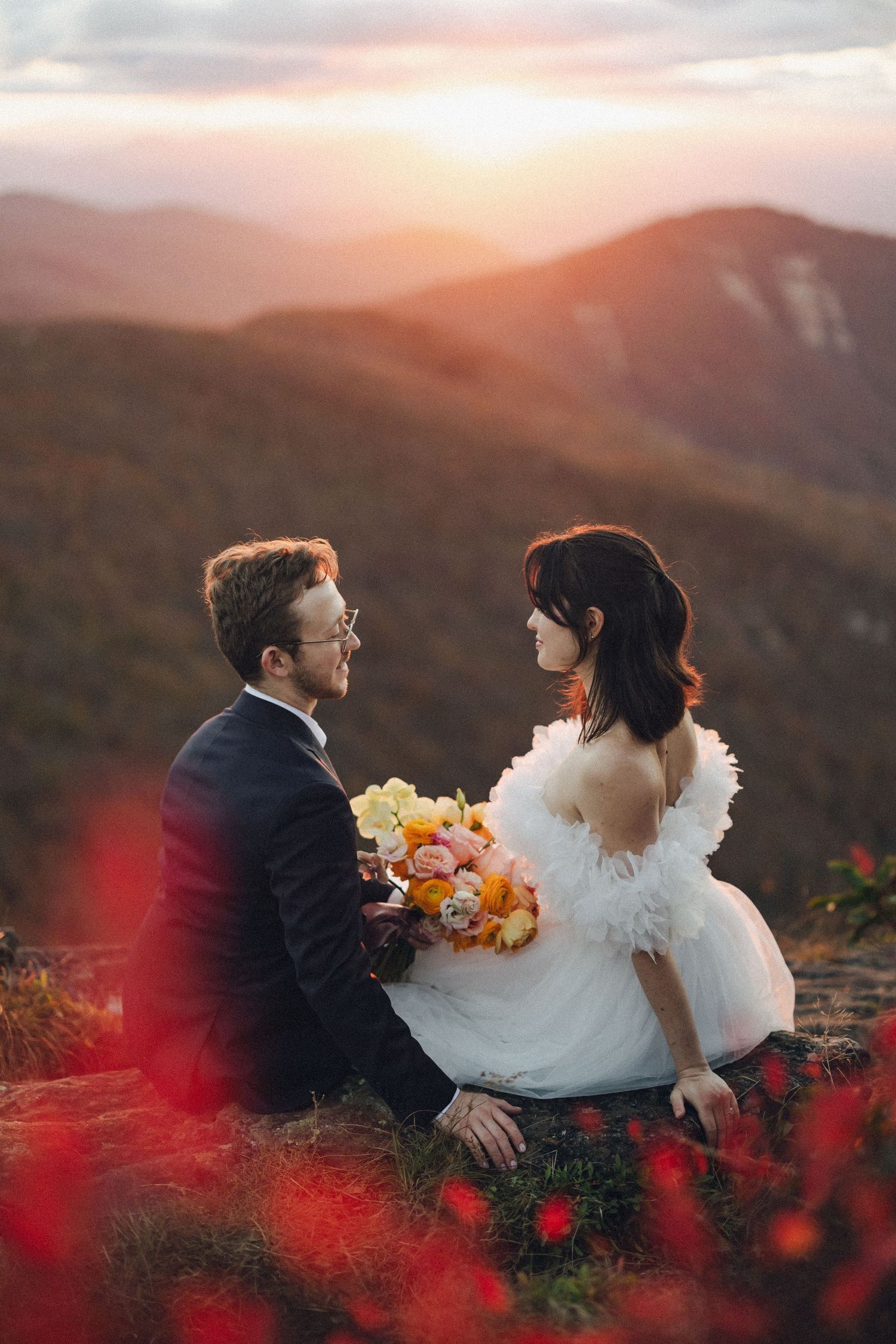 Bride and groom sit facing each other at sunset on a mountain with vibrant red wildflowers in foreground.