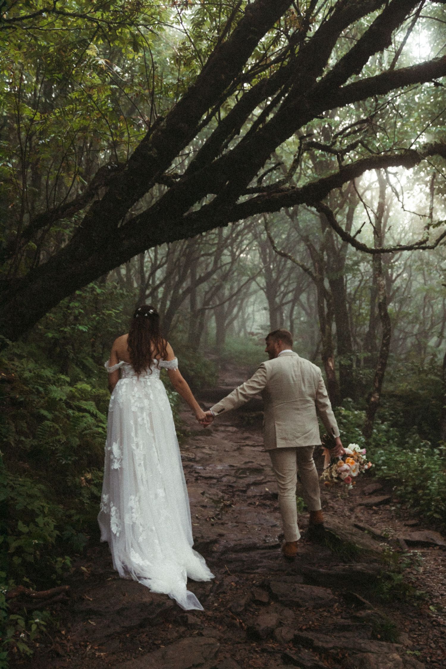 Bride and groom walk hand-in-hand along a misty forest path, she in a floral white gown, he in a beige suit.