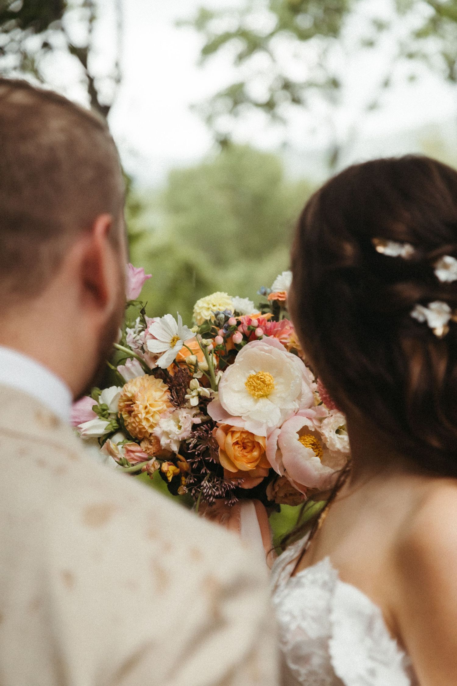 Close-up of bride and groom holding a colorful wedding bouquet with peonies and roses outdoors.