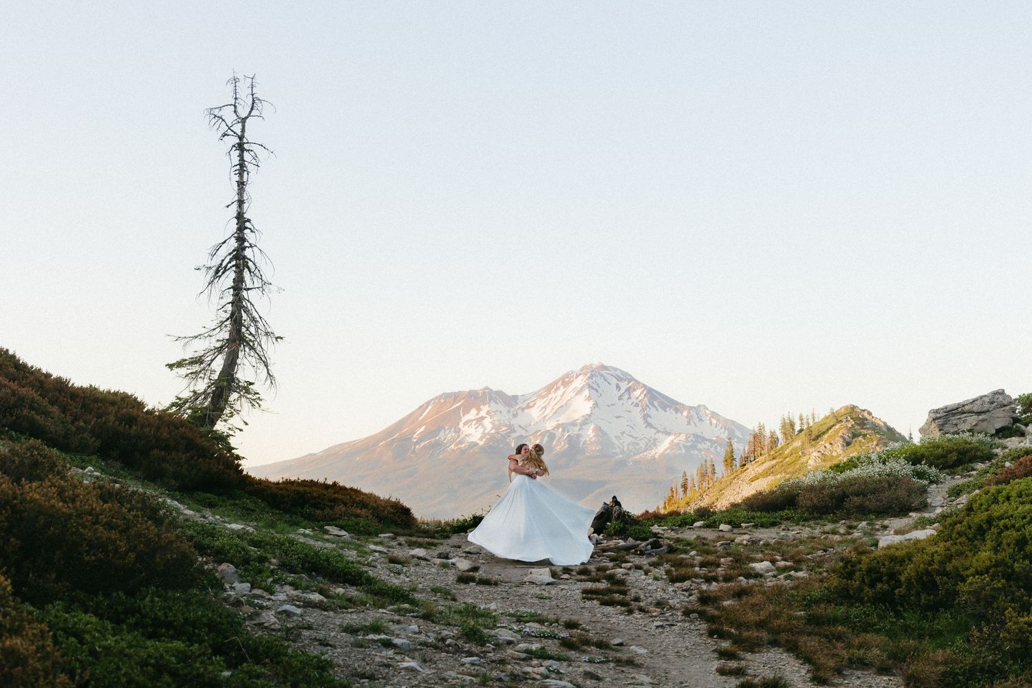 Two brides share a romantic embrace on a mountain trail with a stunning snow-capped peak at sunset.