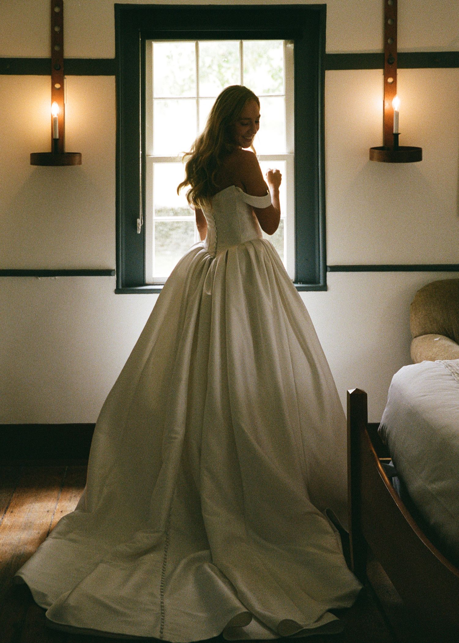 Bride in stunning off-shoulder white ballgown stands by window in soft natural light on her wedding day