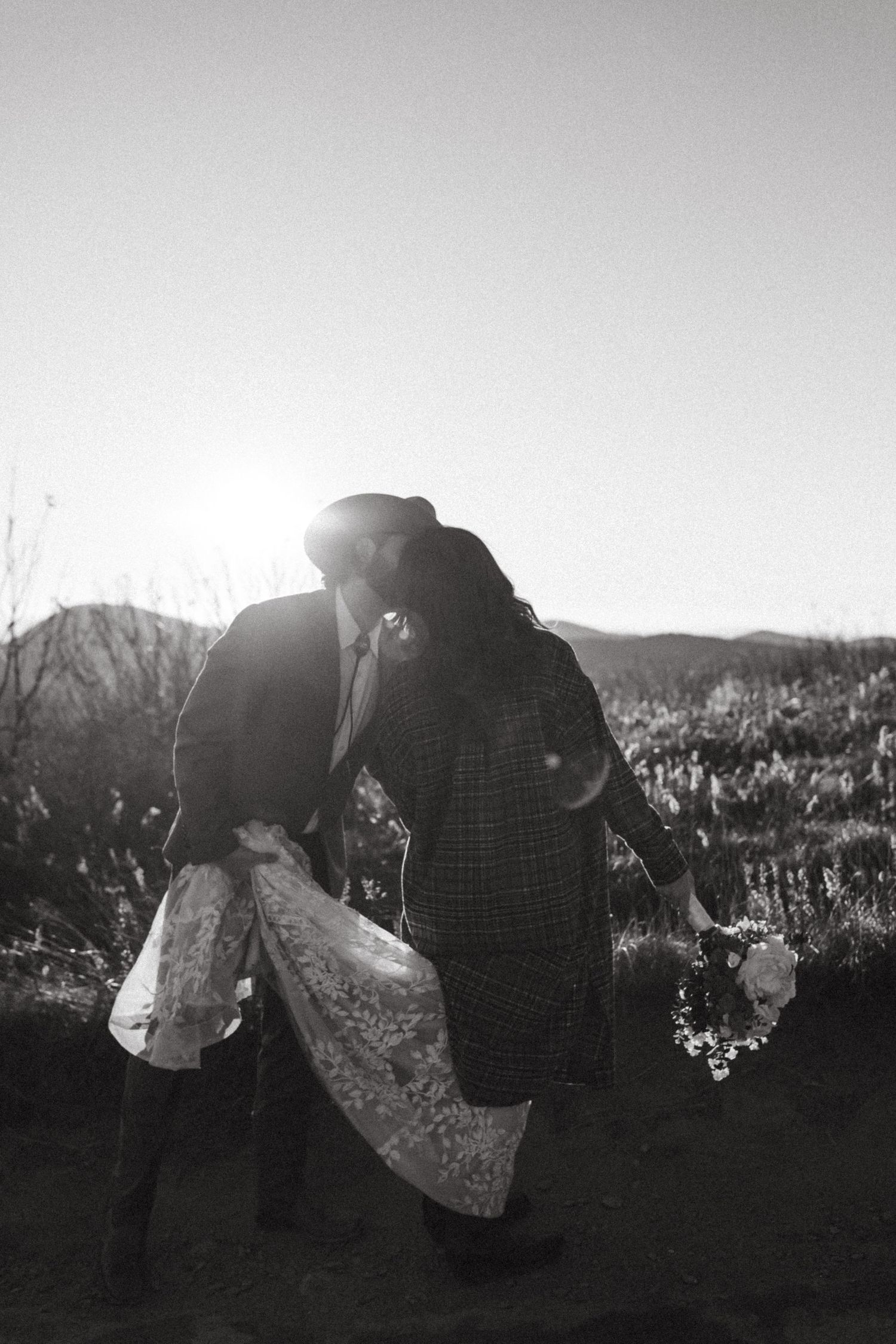 Black and white photo of couple kissing with wedding dress train flowing in mountain landscape.