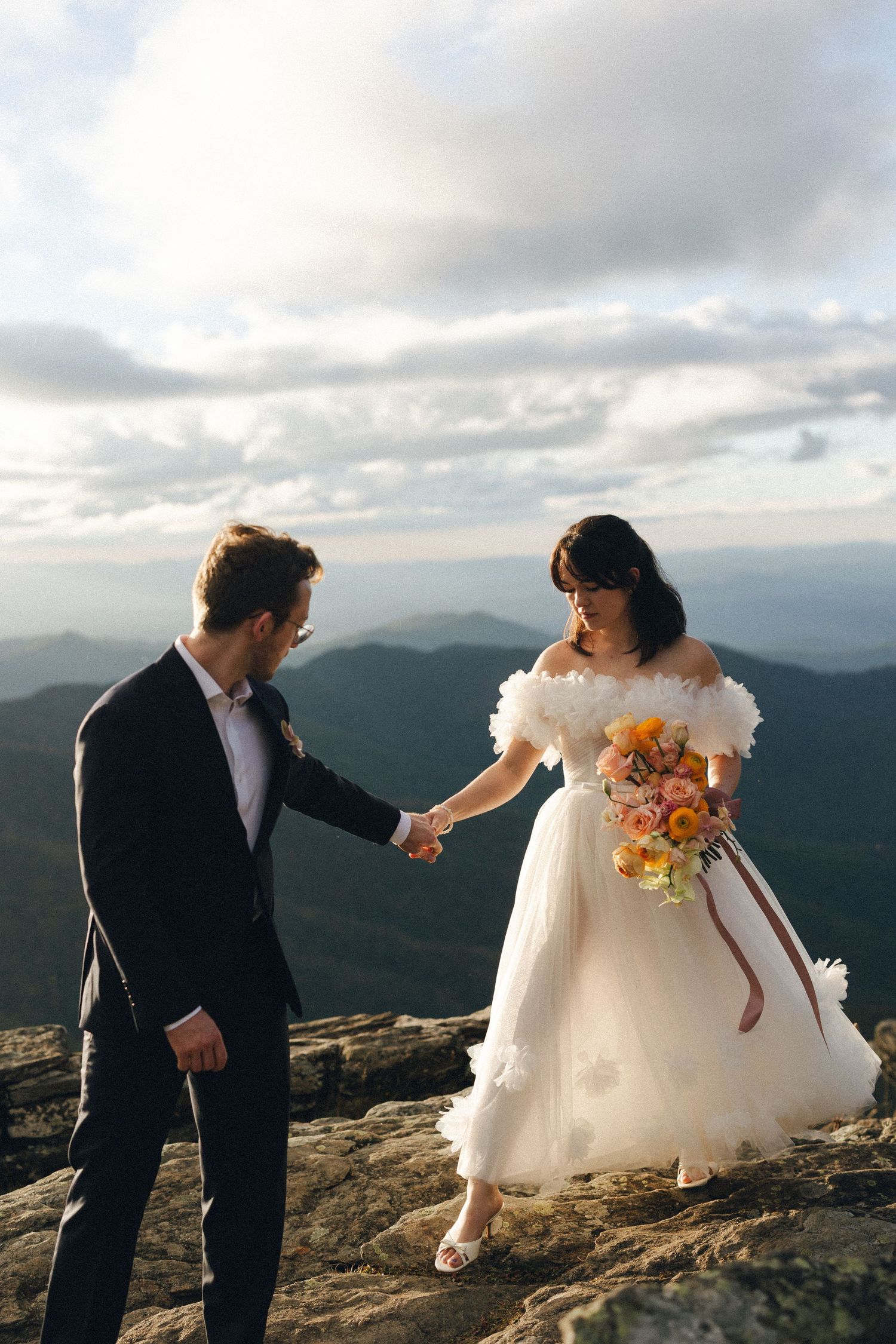 Bride and groom holding hands on a rocky mountain summit with stunning panoramic views behind them.