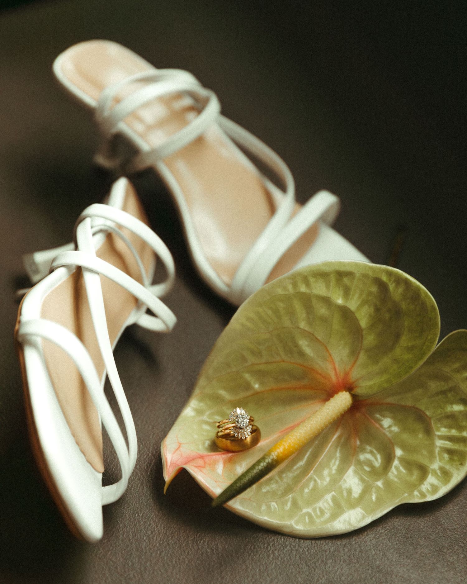 White strappy bridal sandals and a diamond ring resting on a green anthurium leaf on a dark surface.