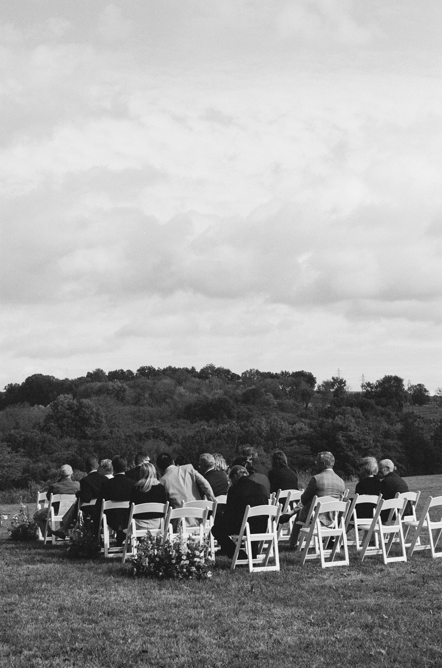 Black and white photo of wedding guests seated in white chairs at outdoor ceremony with scenic countryside backdrop.