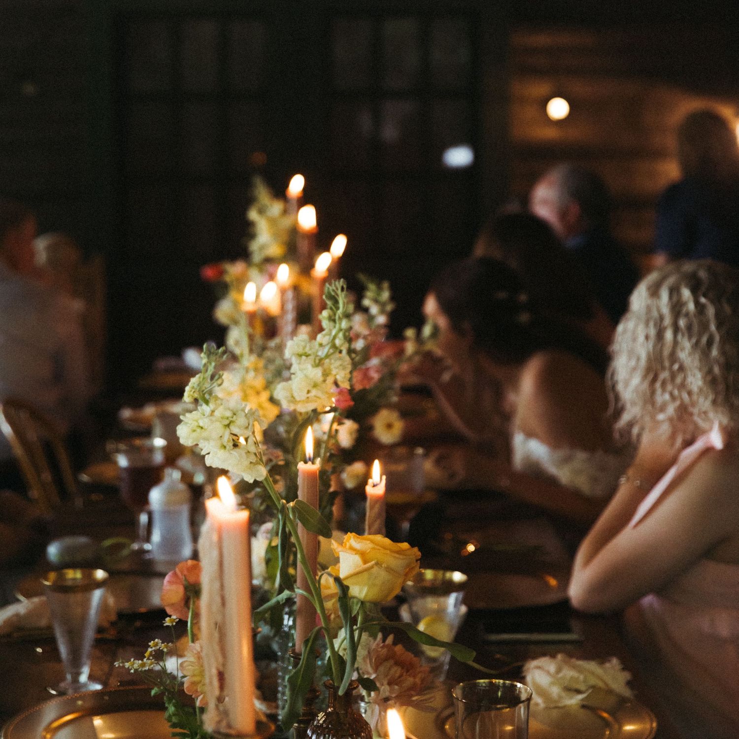 Elegant candlelit wedding reception table with floral centerpieces in a dimly lit rustic barn.