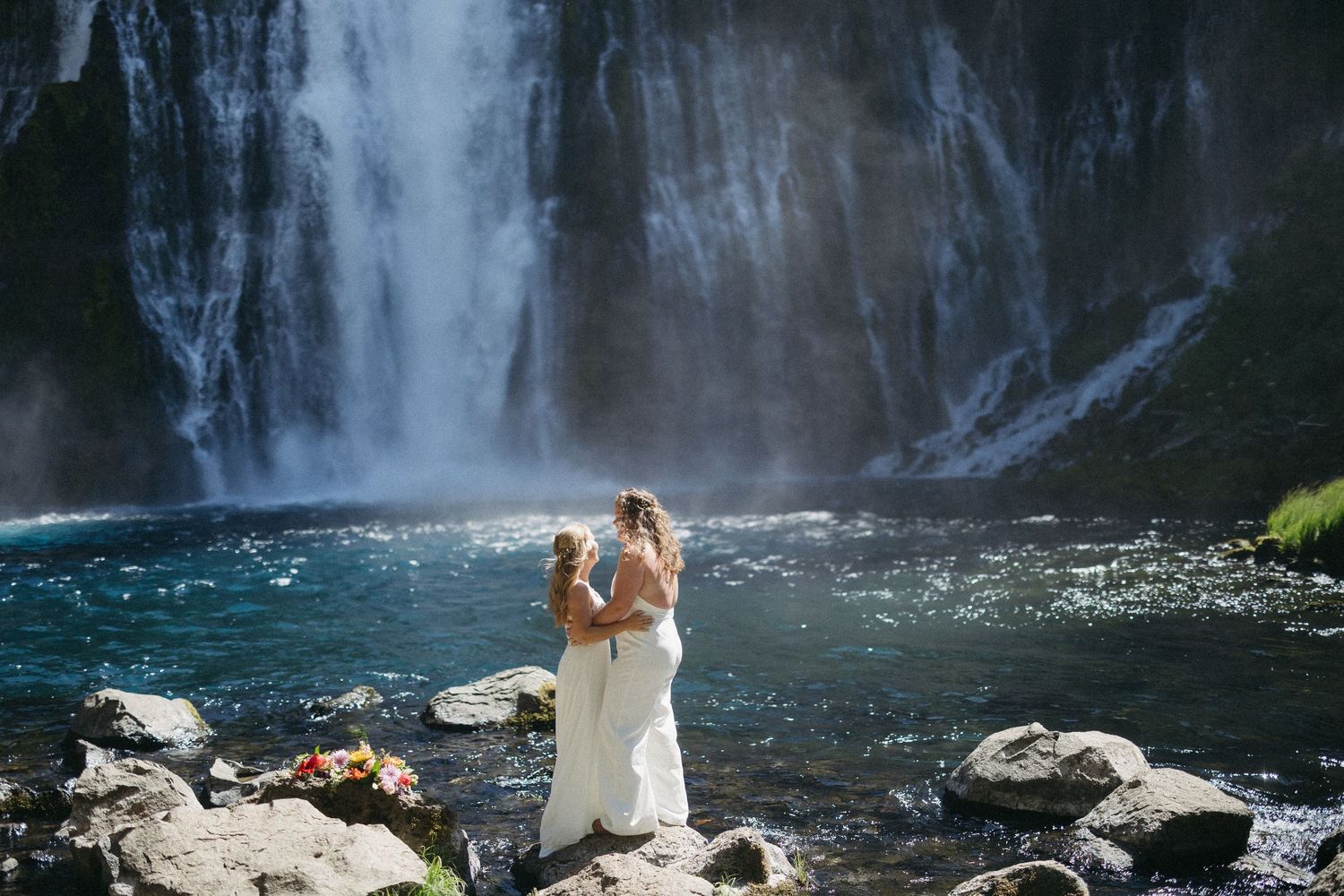 Two brides embrace romantically in front of a breathtaking cascading waterfall with turquoise waters below.