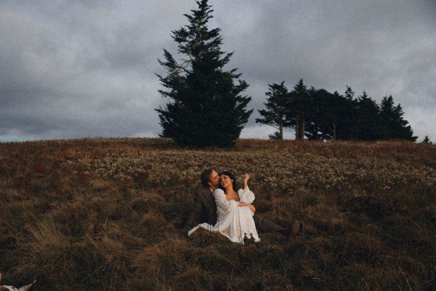 Two brides laugh and embrace joyfully in an open meadow with evergreen trees under an overcast sky.