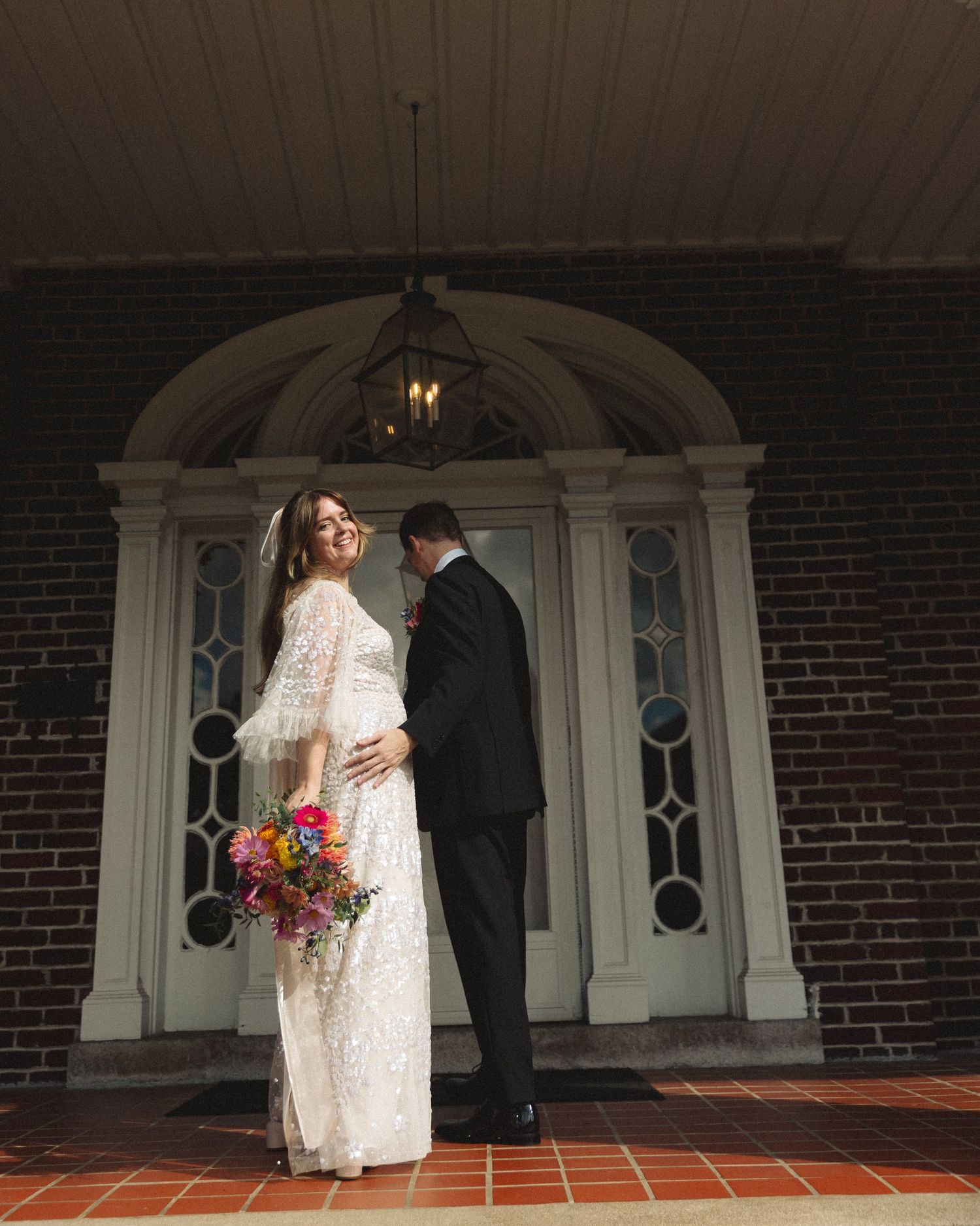 Smiling bride in lace gown poses with groom at arched brick church entrance, holding a bright multicolored bouquet.