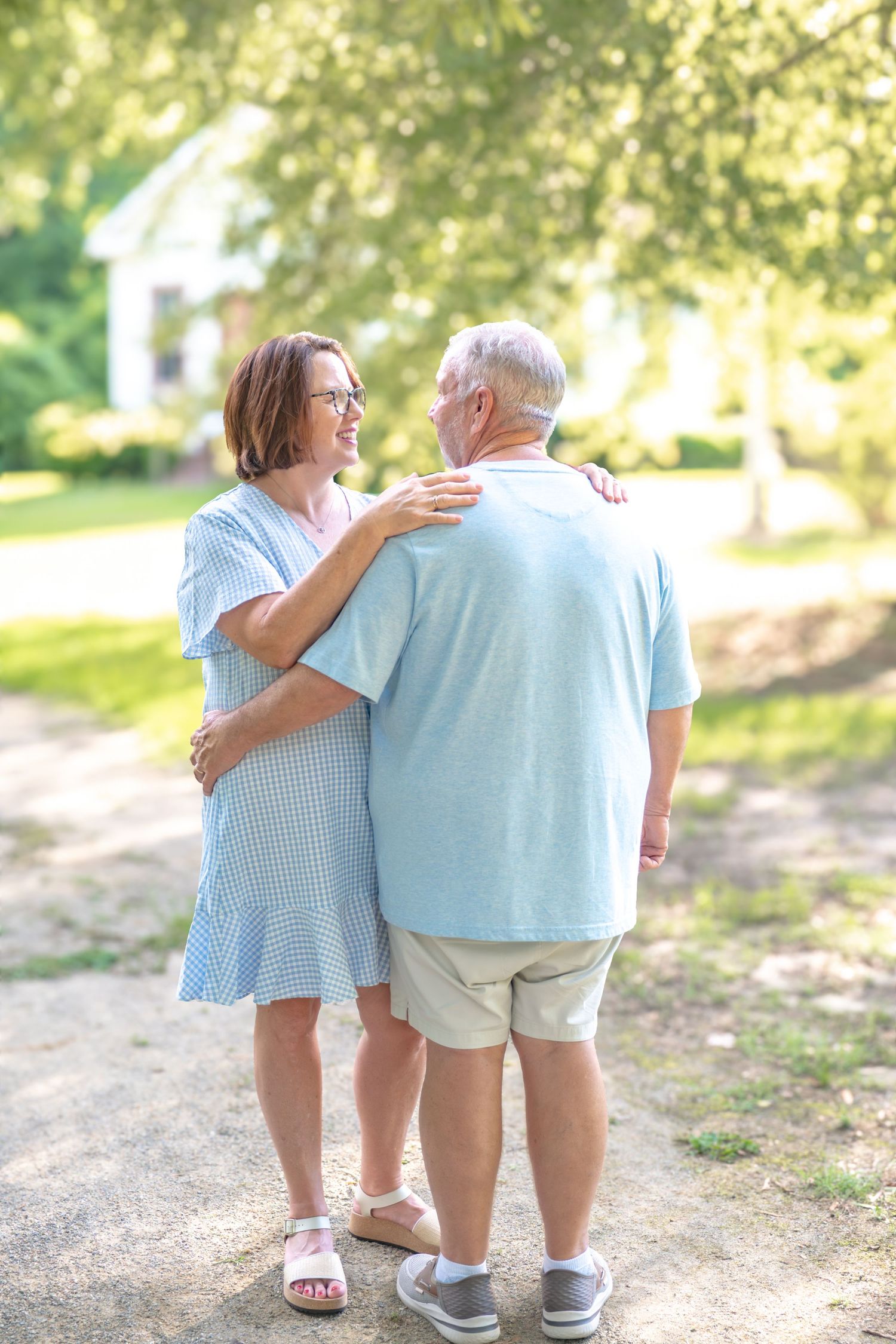 Older couple embracing and smiling at each other while walking on a sunny tree-lined path.