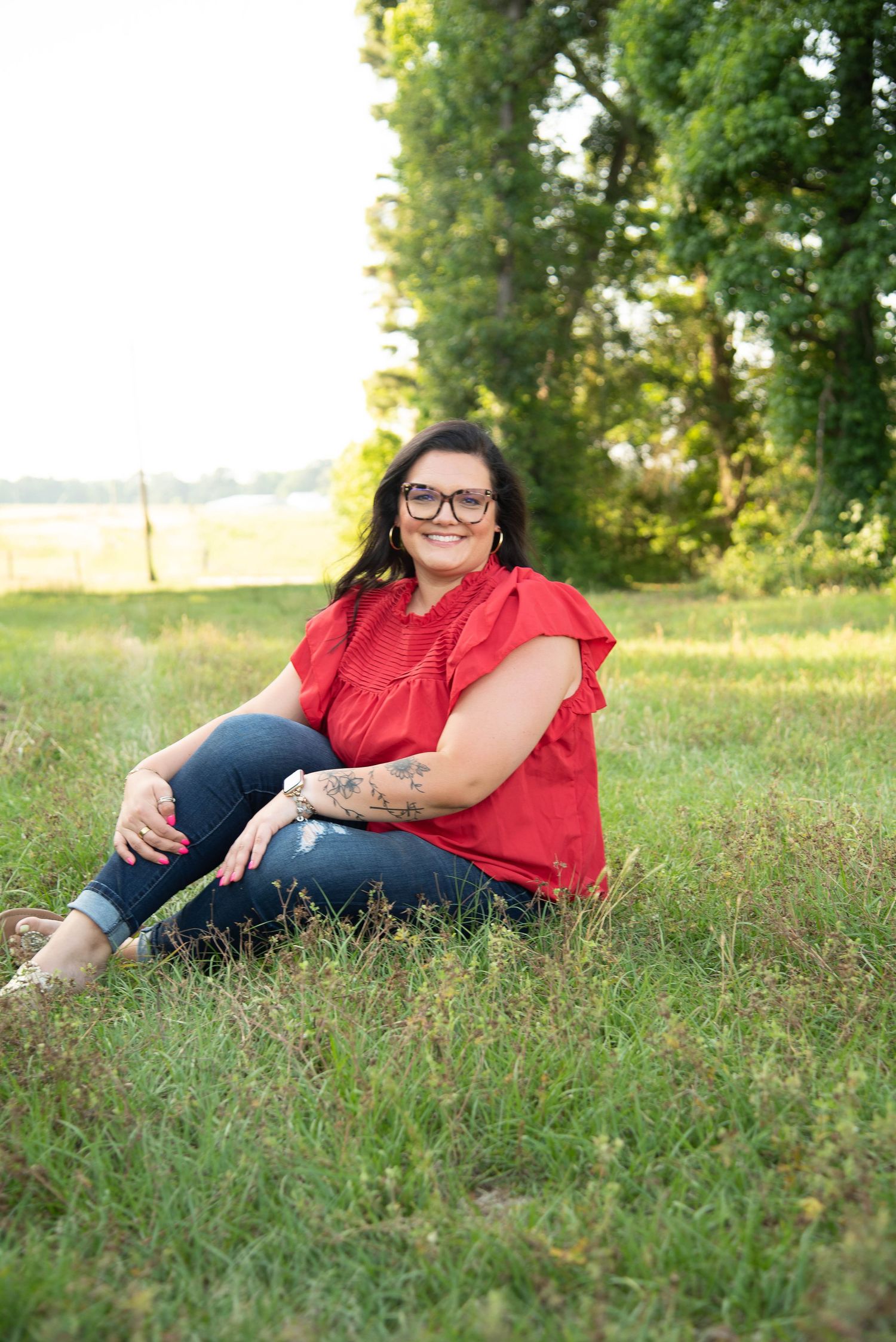 Woman in red top and jeans sitting in green grass field smiling outdoors.