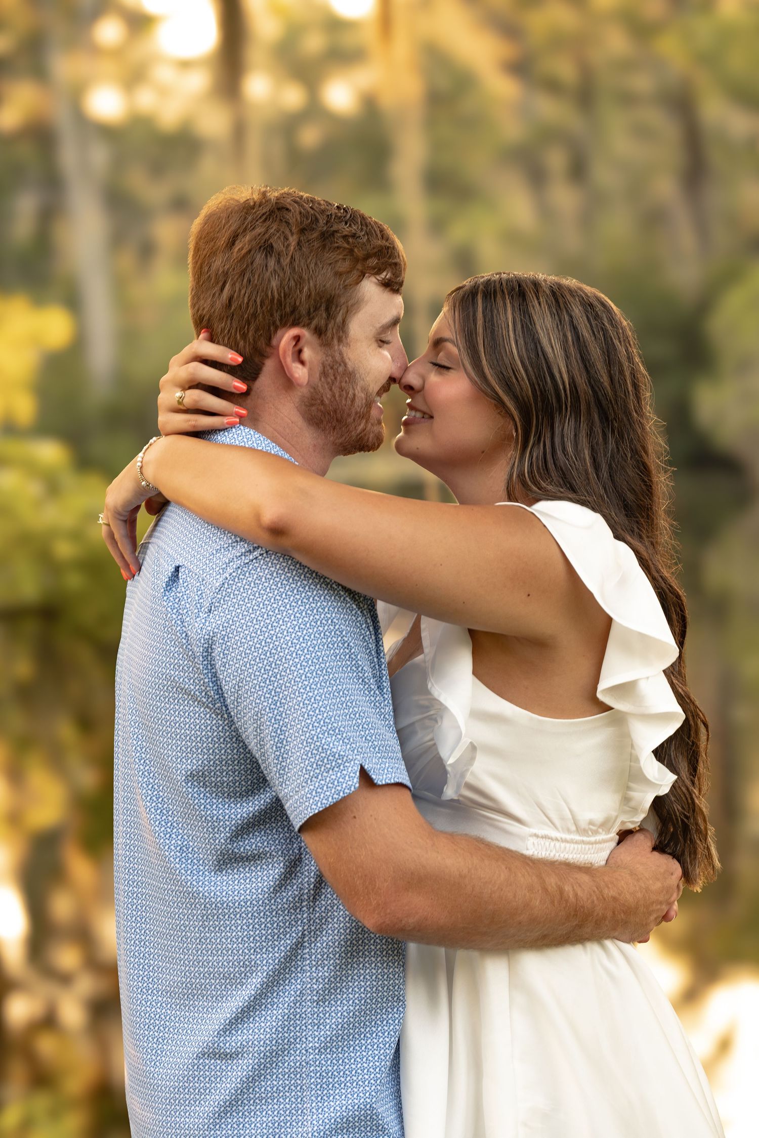 Couple in romantic embrace outdoors, woman in white dress and man in blue shirt, sharing a tender moment near trees.