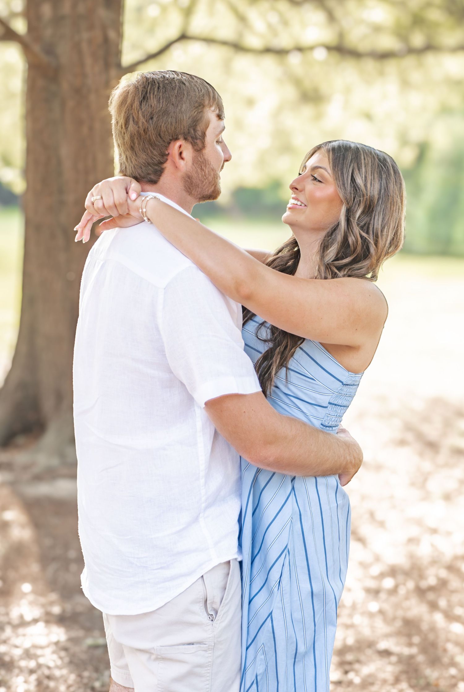 Couple embracing and smiling at each other near a large tree in a sun-dappled outdoor setting.