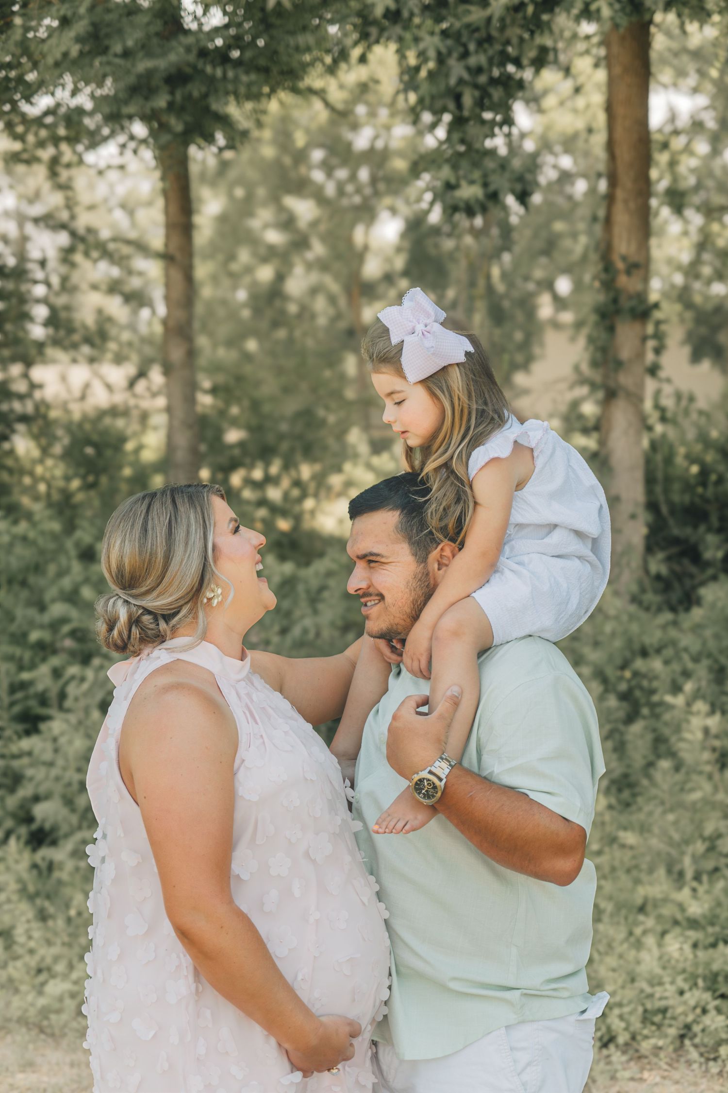A pregnant woman in white dress smiles with man carrying young girl on shoulders outdoors.