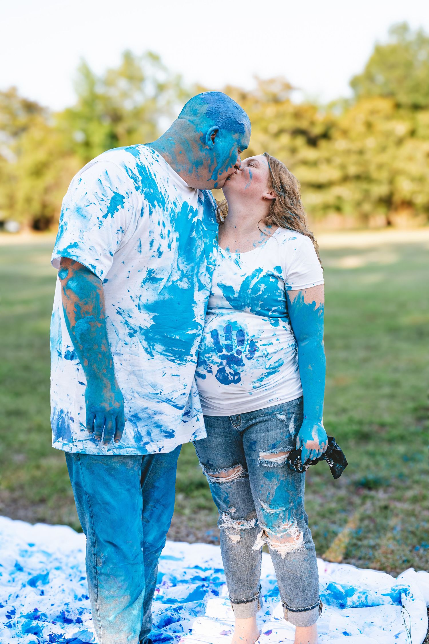 Couple kissing outdoors covered in blue paint during a gender reveal celebration, standing on white sheet in park.