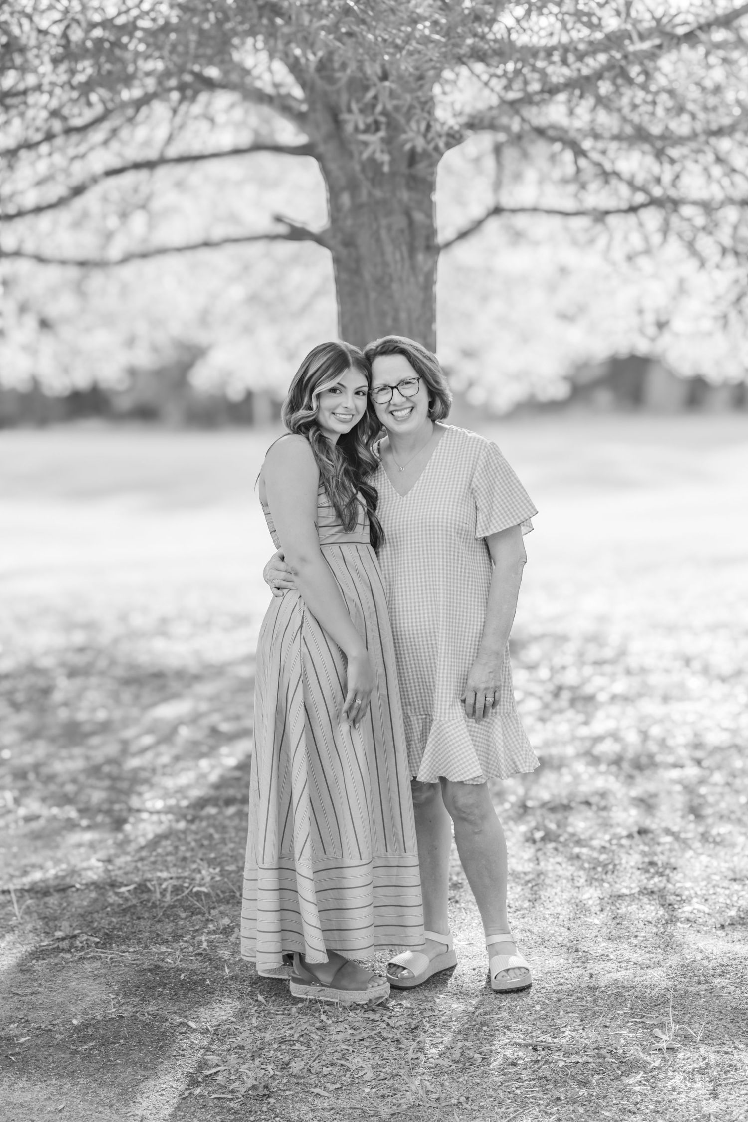 Black and white portrait of two women hugging and smiling together under a large tree outdoors.