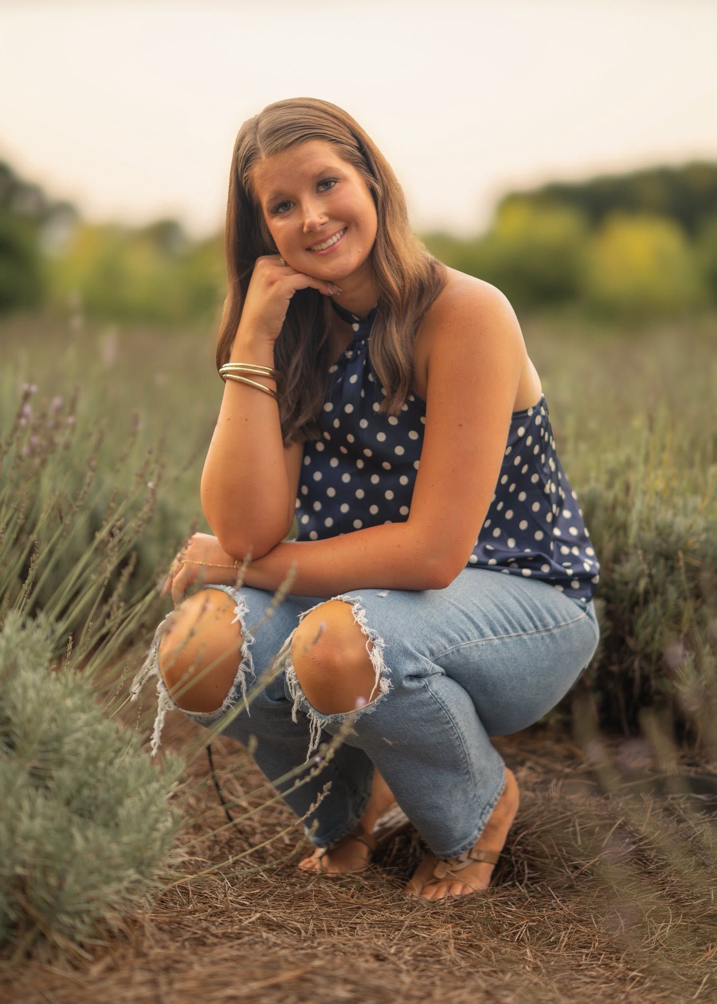 Young woman in polka dot top and distressed jeans crouching in a lavender field, smiling warmly.