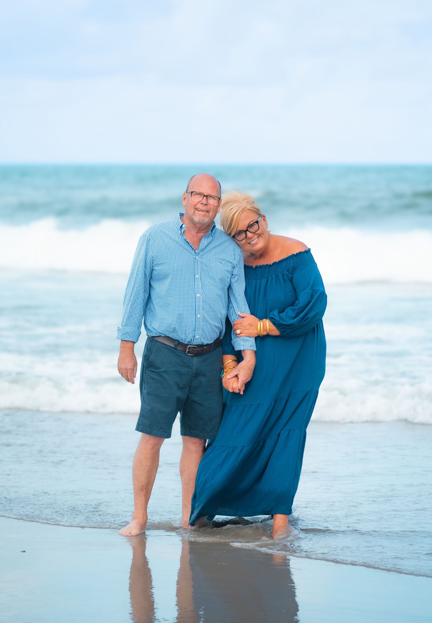 Couple smiling and holding hands on a beach, wearing blue outfits, with ocean waves in the background.