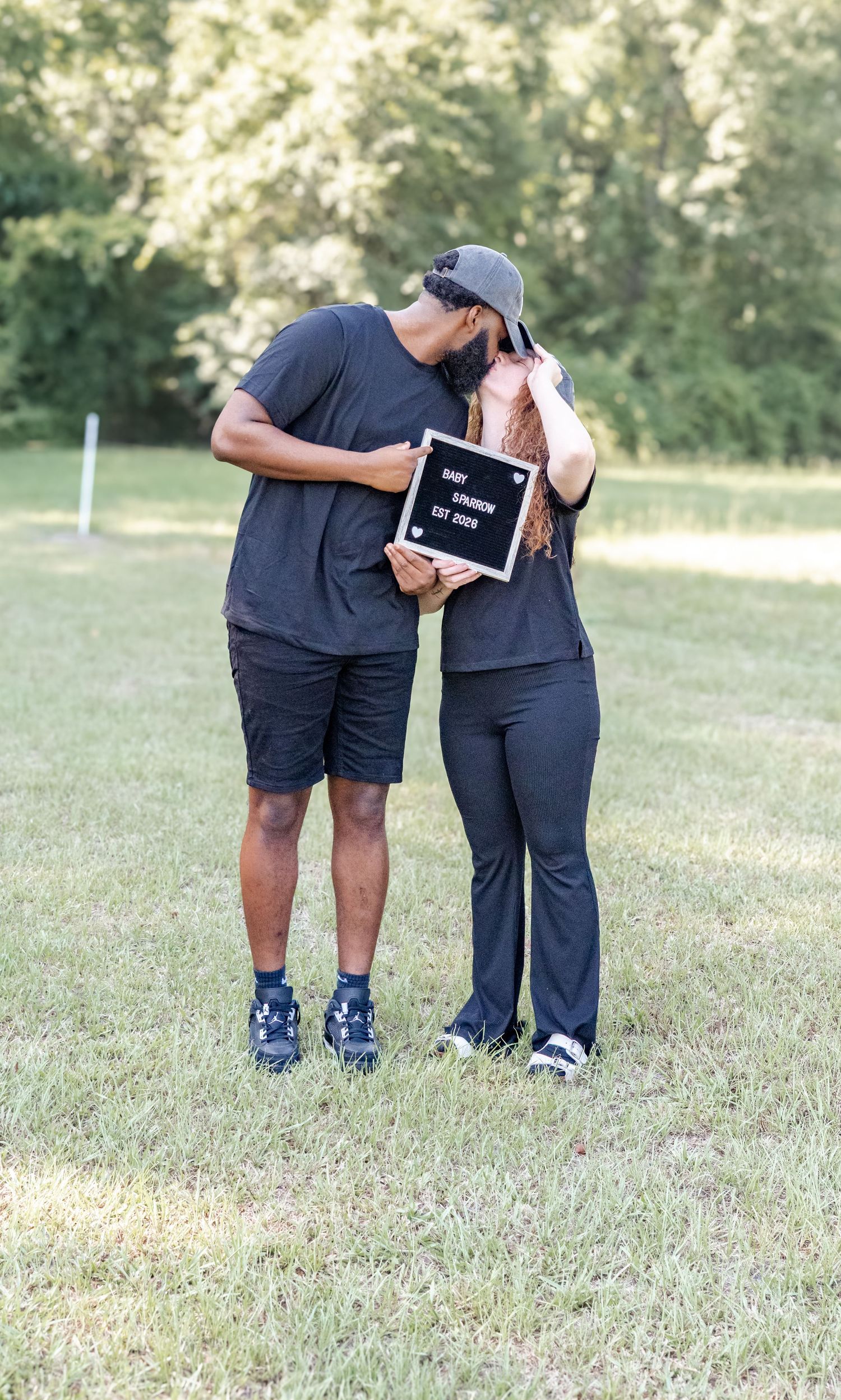 Couple kissing outdoors holding a Baby Sparrow Est 2026 letterboard sign in a grassy park setting.