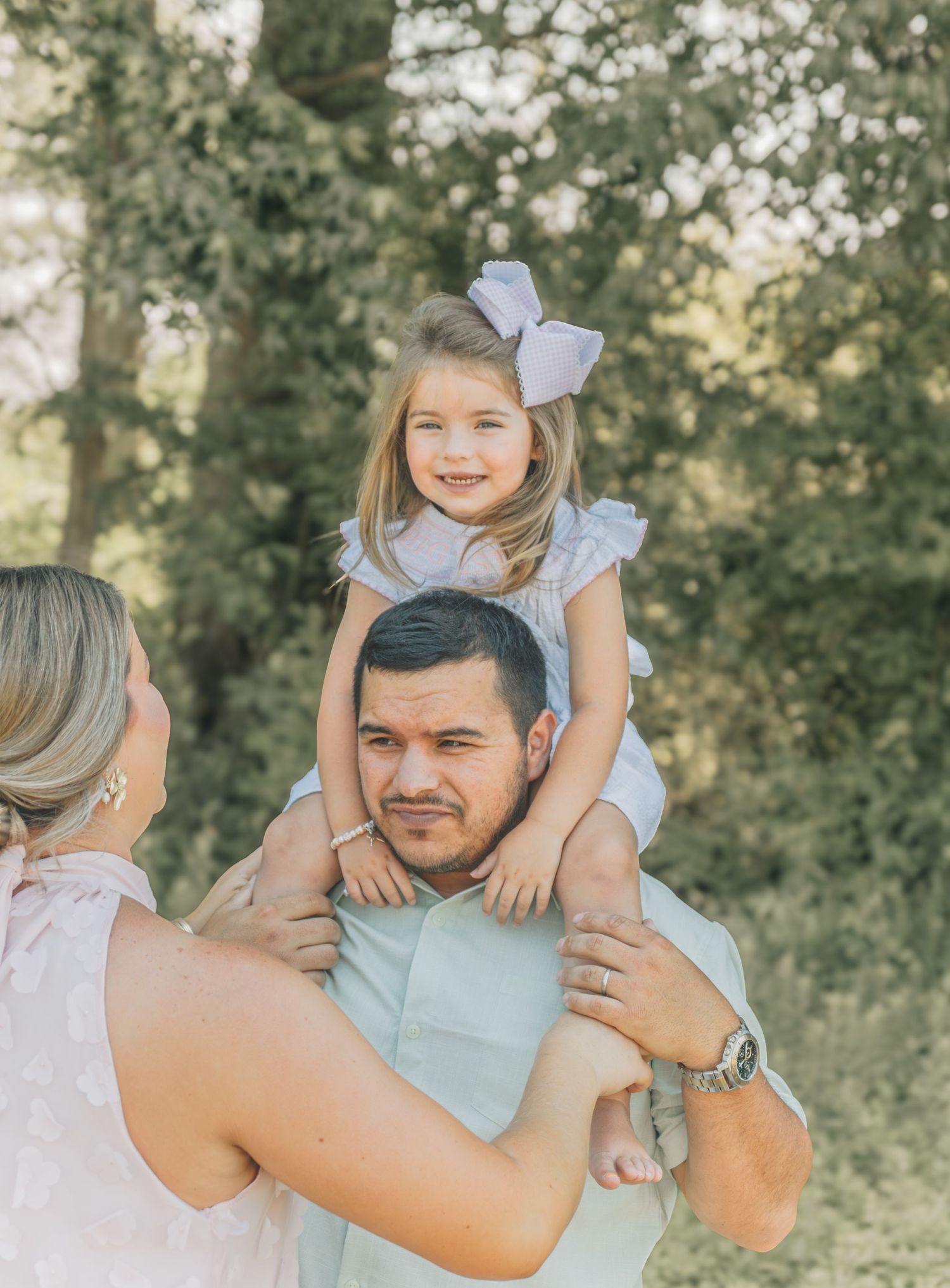 A young girl with a white bow sits on a man's shoulders while a woman stands beside them outdoors.