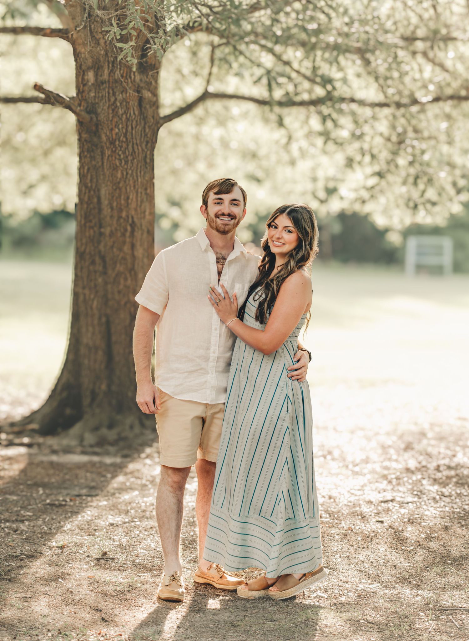 Couple posing under a large tree in a park, man in beige shorts and shirt, woman in a striped maxi dress.