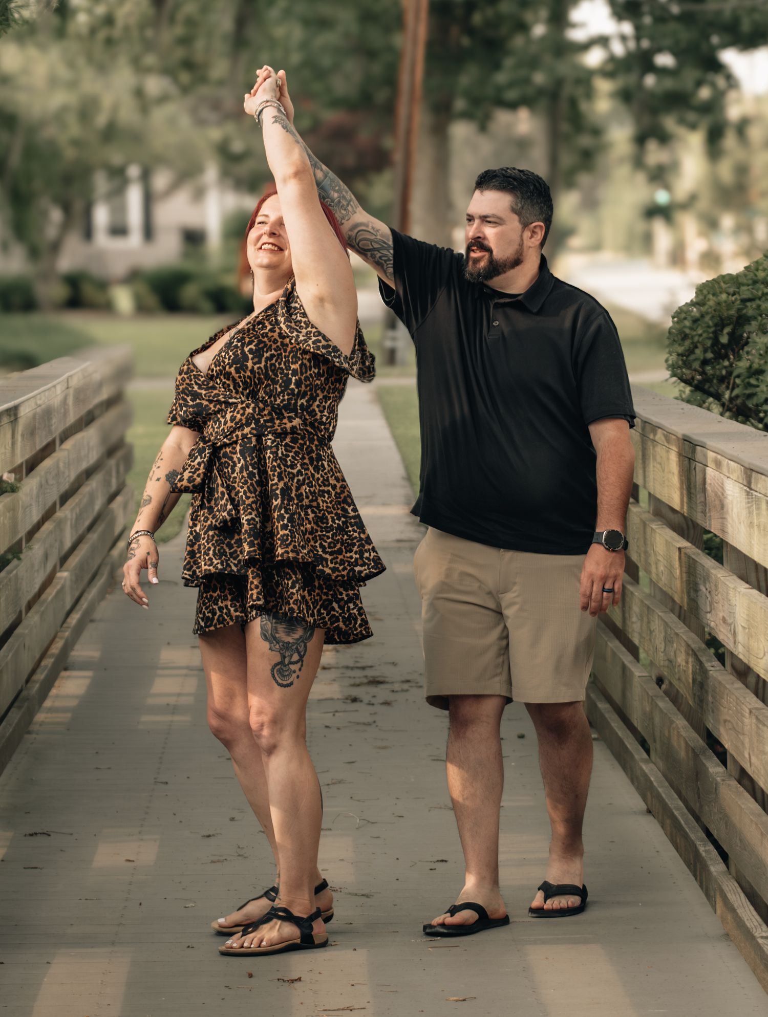 A couple dances on a tree-lined walkway, man in black shirt spinning woman in leopard print dress.