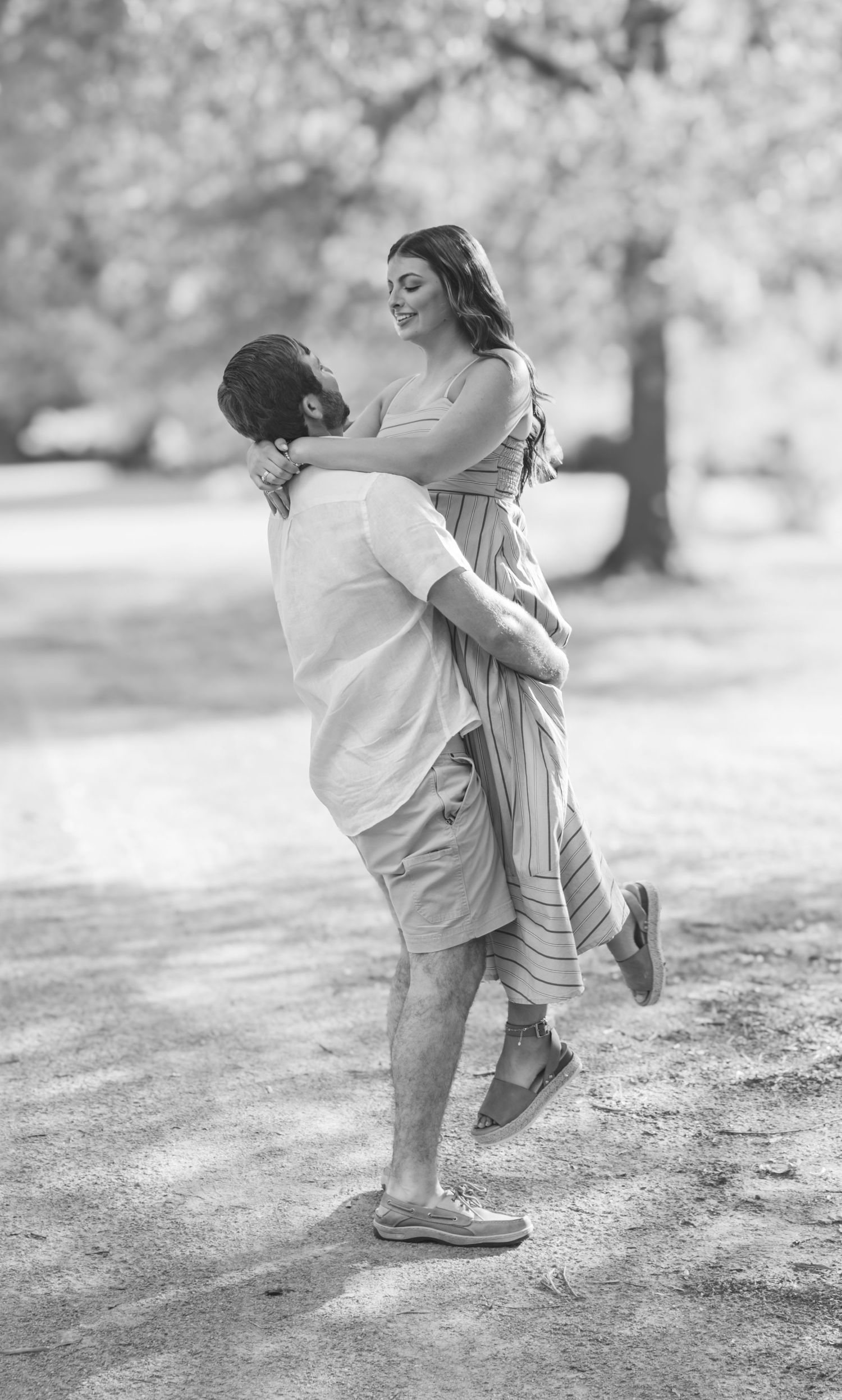Black and white photo of a man lifting a smiling woman in a park, sharing a joyful romantic moment outdoors.