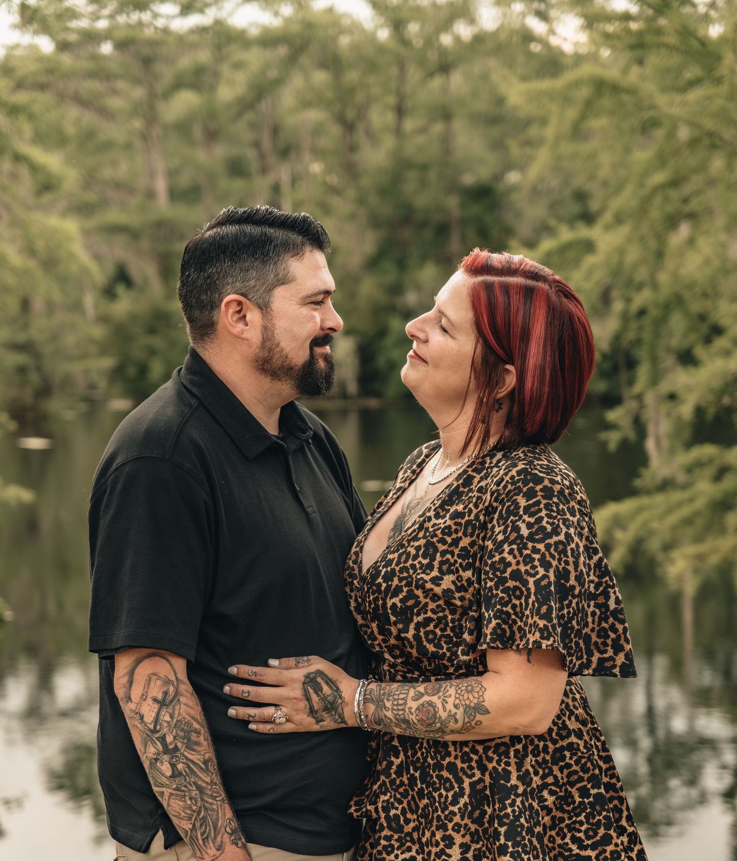Tattooed couple in a romantic embrace outdoors near a lake, wearing black and leopard print outfits.