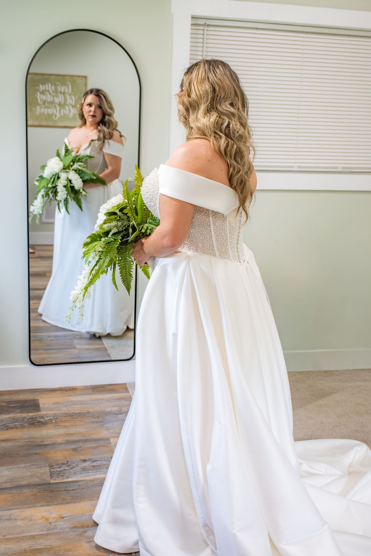 Bride in off-shoulder white gown holding green bouquet, viewed from behind reflecting in arched mirror.