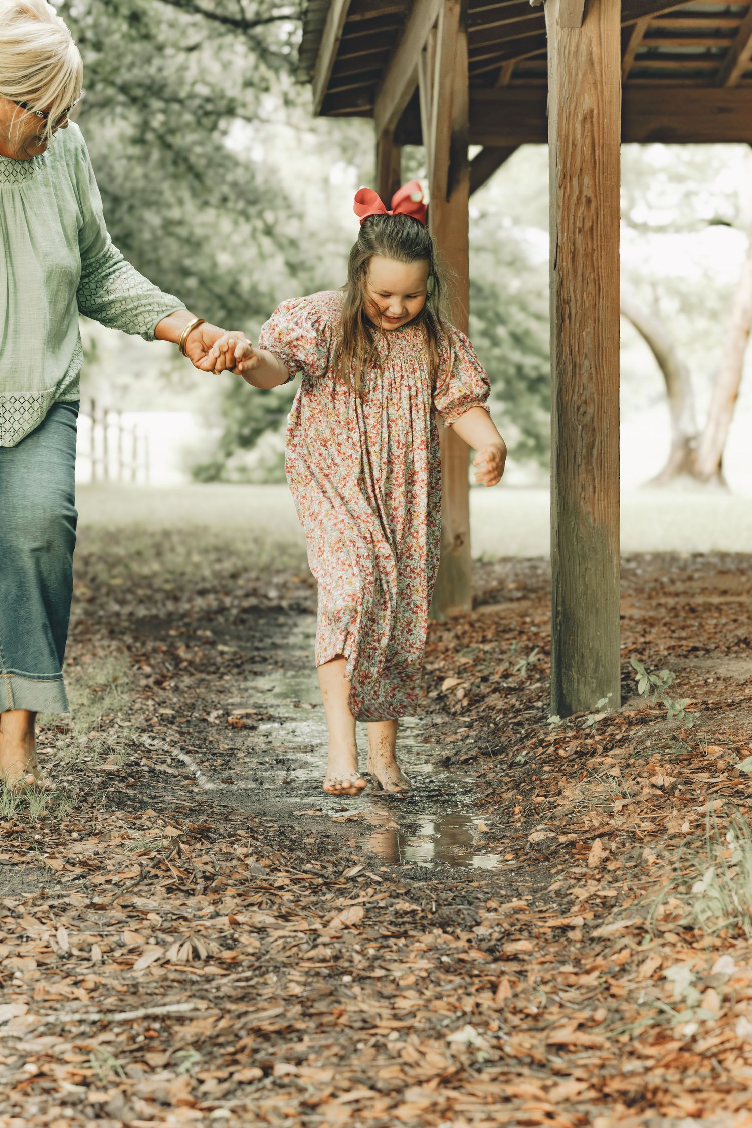 A young girl in a floral dress splashes in puddles while holding hands with an adult outdoors.
