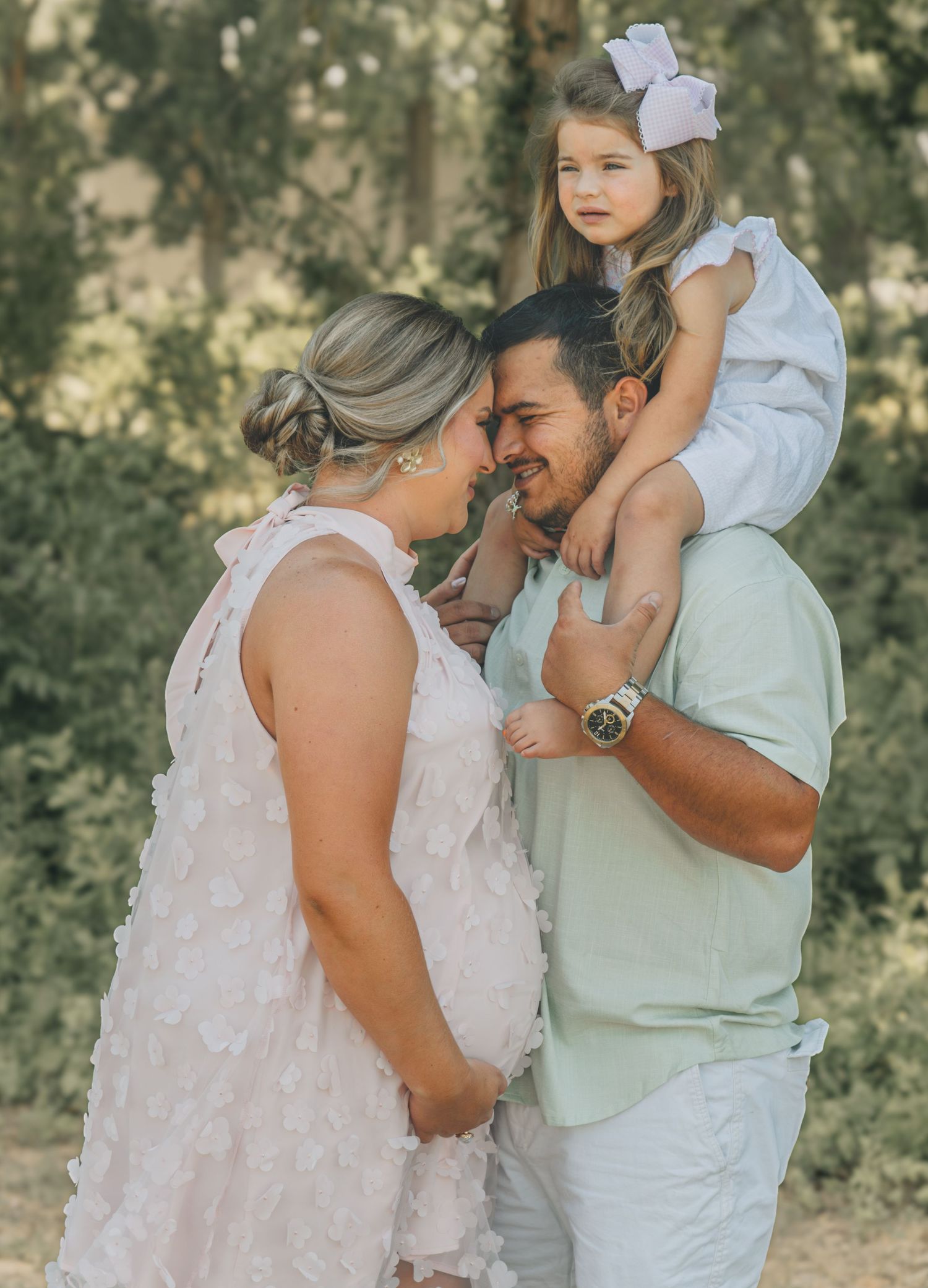 A happy family poses outdoors, with a man holding a little girl on his shoulders while facing a woman in a white floral dress.