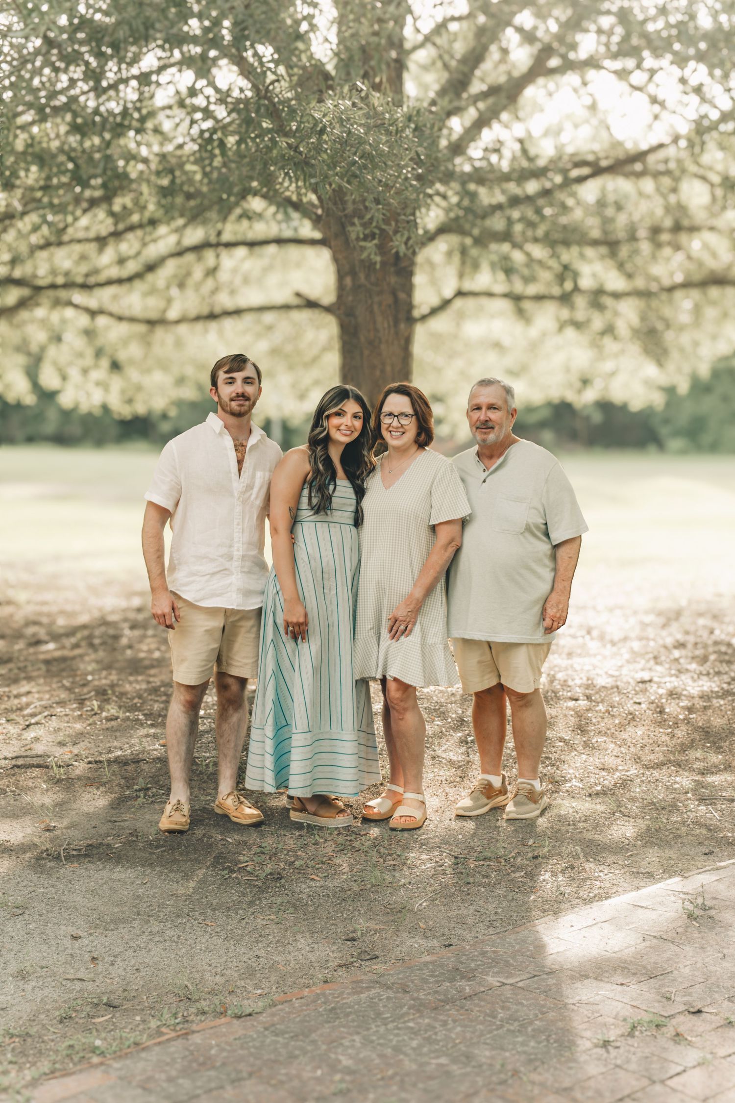 Family of four smiling together under a large tree in a sunny outdoor park setting.