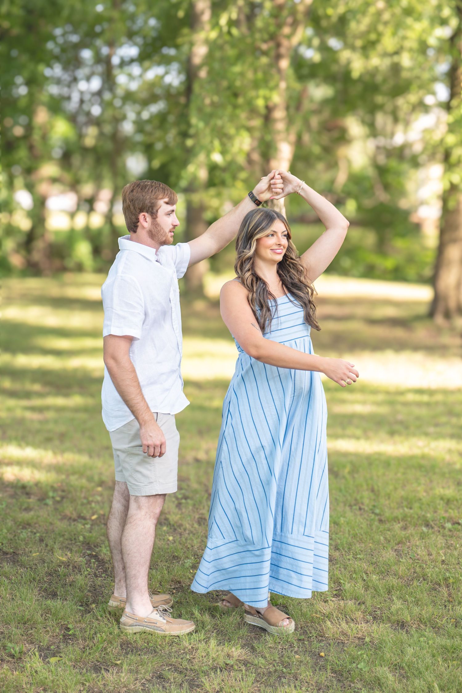 Man spinning smiling woman in blue striped maxi dress in a sunlit green park setting.