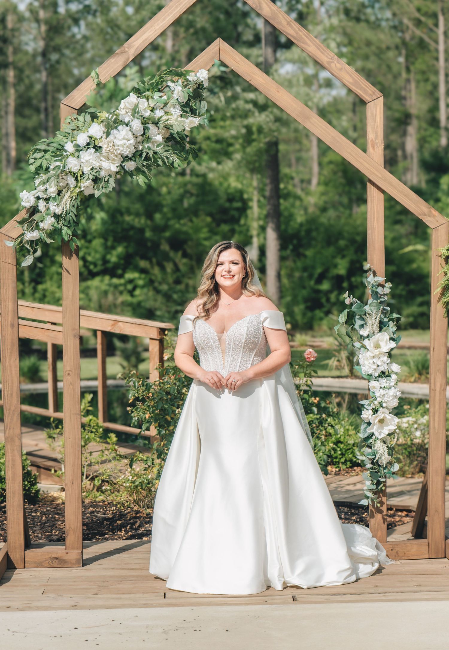 Bride in off-shoulder ball gown stands under wooden arch with white florals at outdoor wedding ceremony.