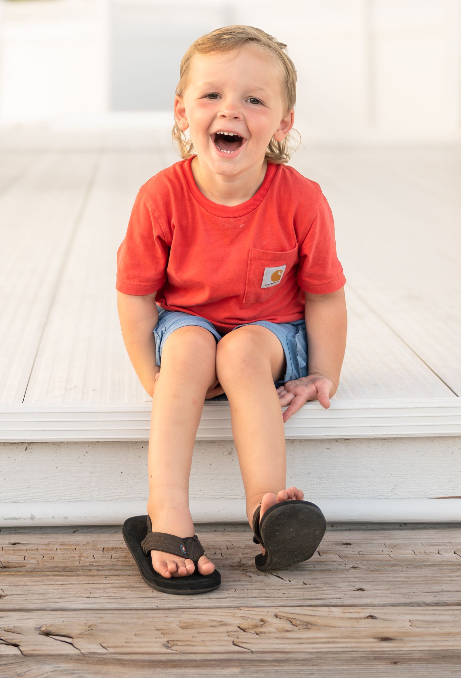 Laughing toddler boy in red Carhartt shirt and denim shorts sitting on white wooden steps wearing flip flops.