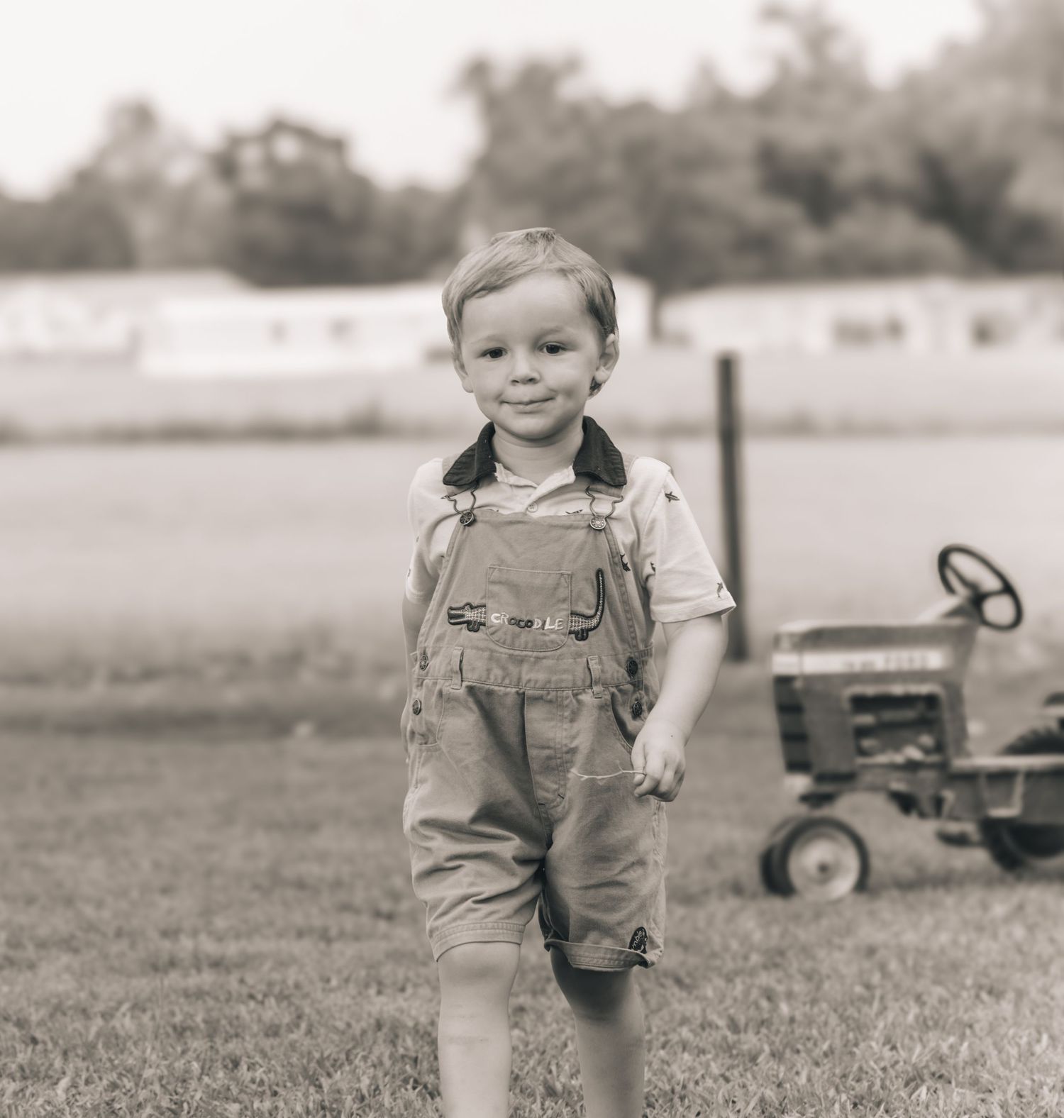 Young boy in overalls smiling on a farm with a tractor in the background, vintage black and white photo.